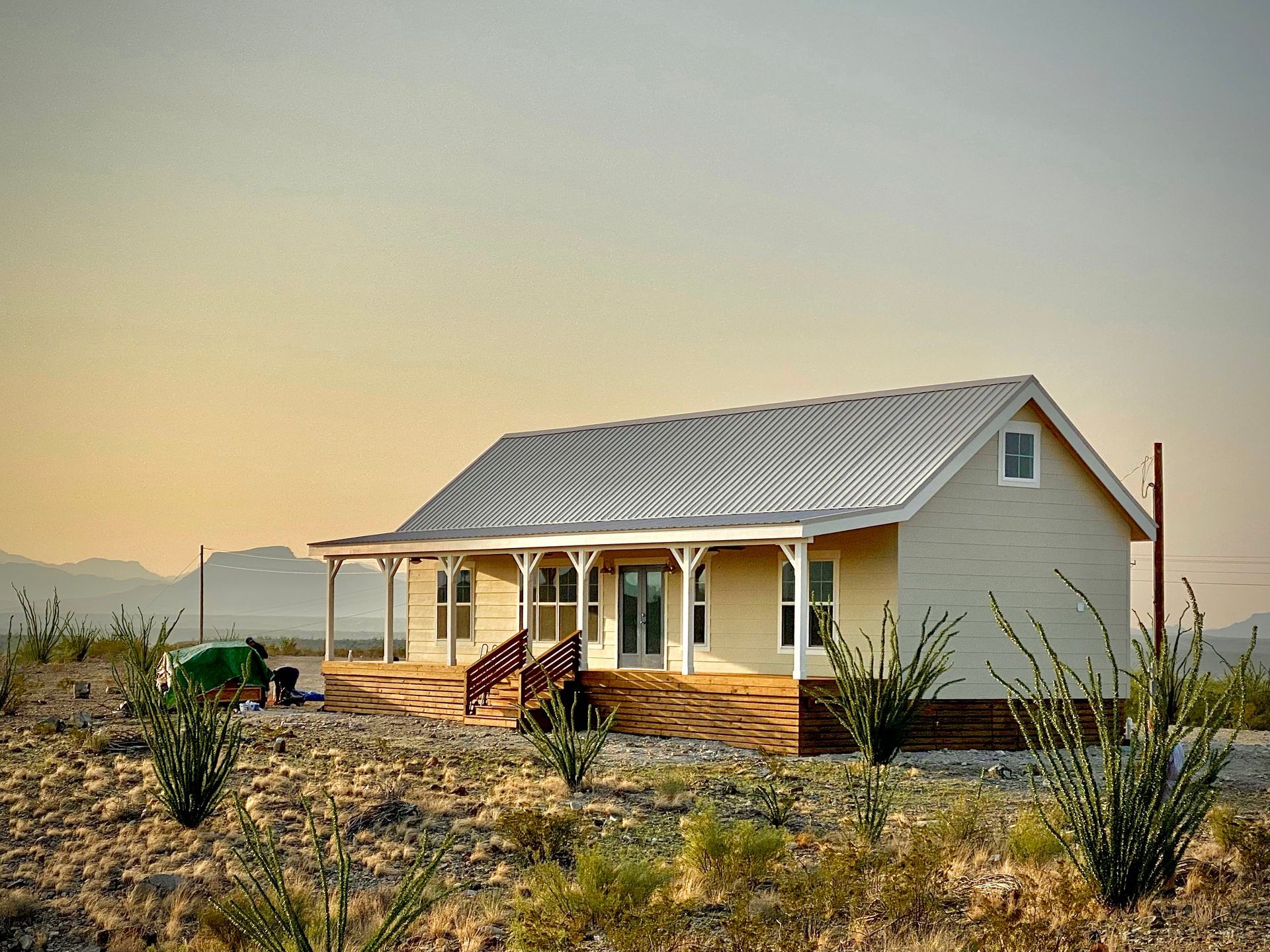 Light-colored house with porch, metal roof, and surrounding desert vegetation; clear sky.