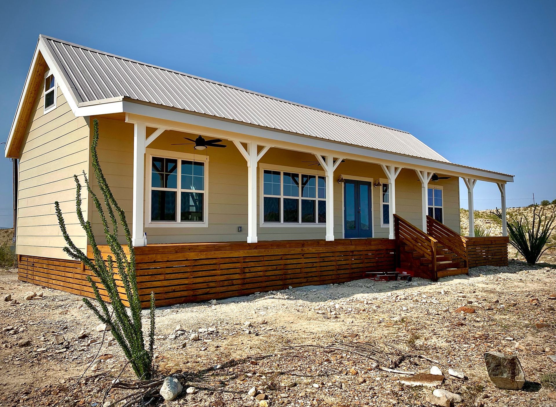 Beige cabin with porch, metal roof, and wooden deck in a desert landscape under a clear blue sky.