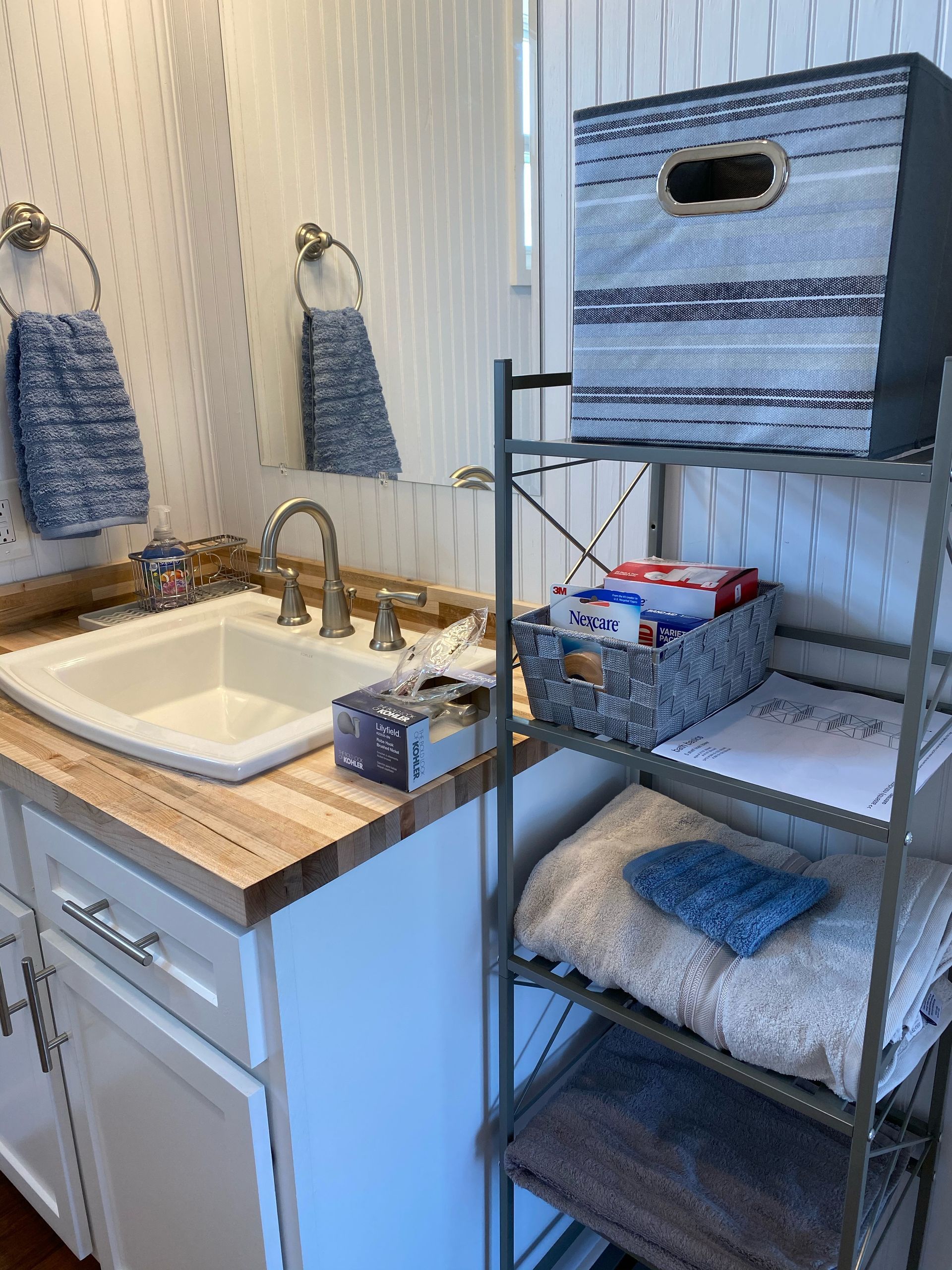 Bathroom with white sink, blue towels, and storage shelves. A blue-striped box is on top shelf.