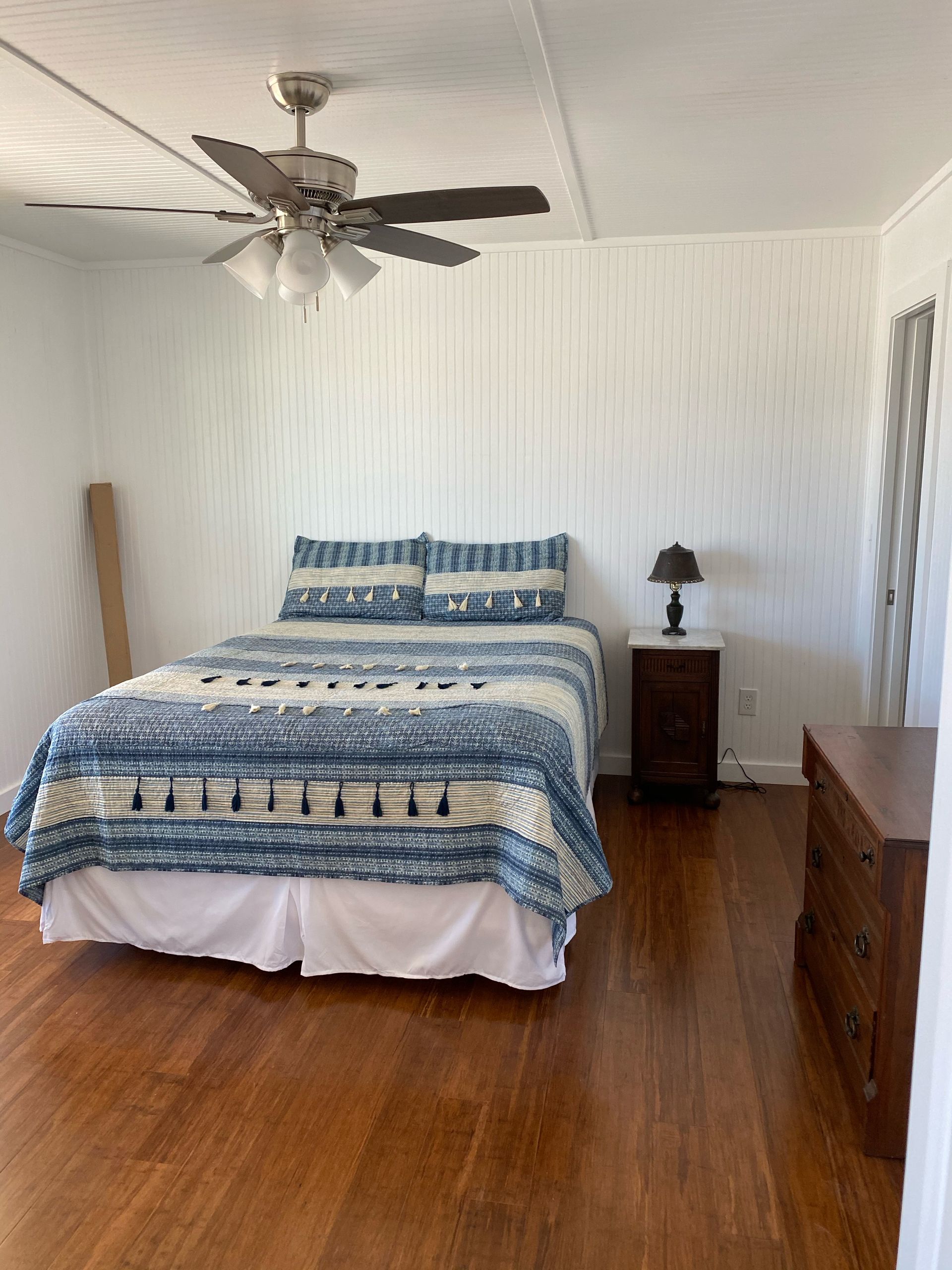 Bedroom with wooden floor, bed with blue and white quilt, dark nightstand, and dresser. Ceiling fan.