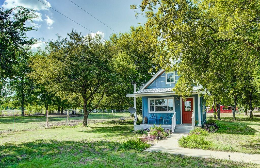 Blue cottage with red door, surrounded by trees and a green lawn under a sunny sky.