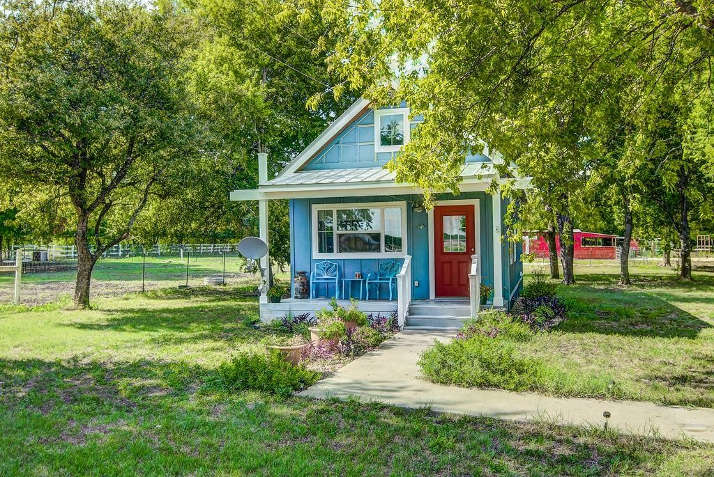 Blue cottage with red door, surrounded by trees and a grassy yard.