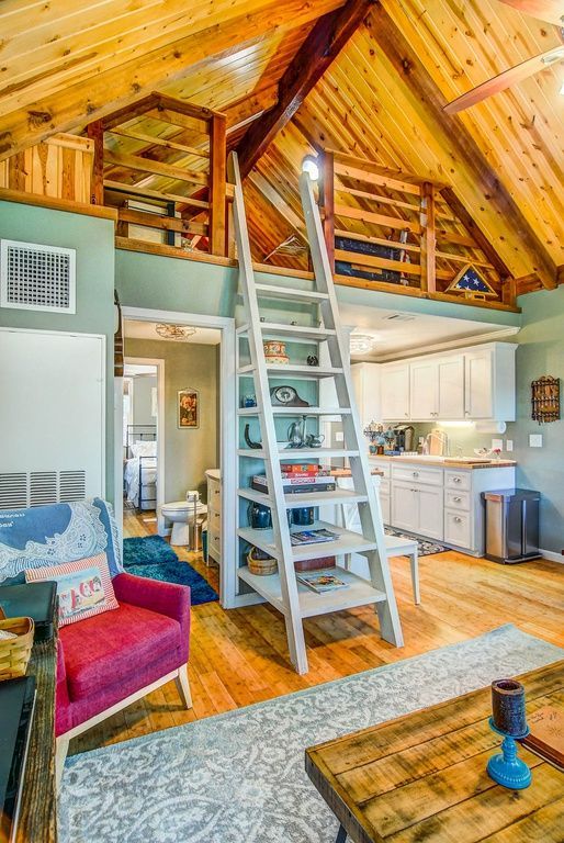 Cozy cabin interior with loft, ladder, and wood beams.  Red chair, kitchen, and blue rug.