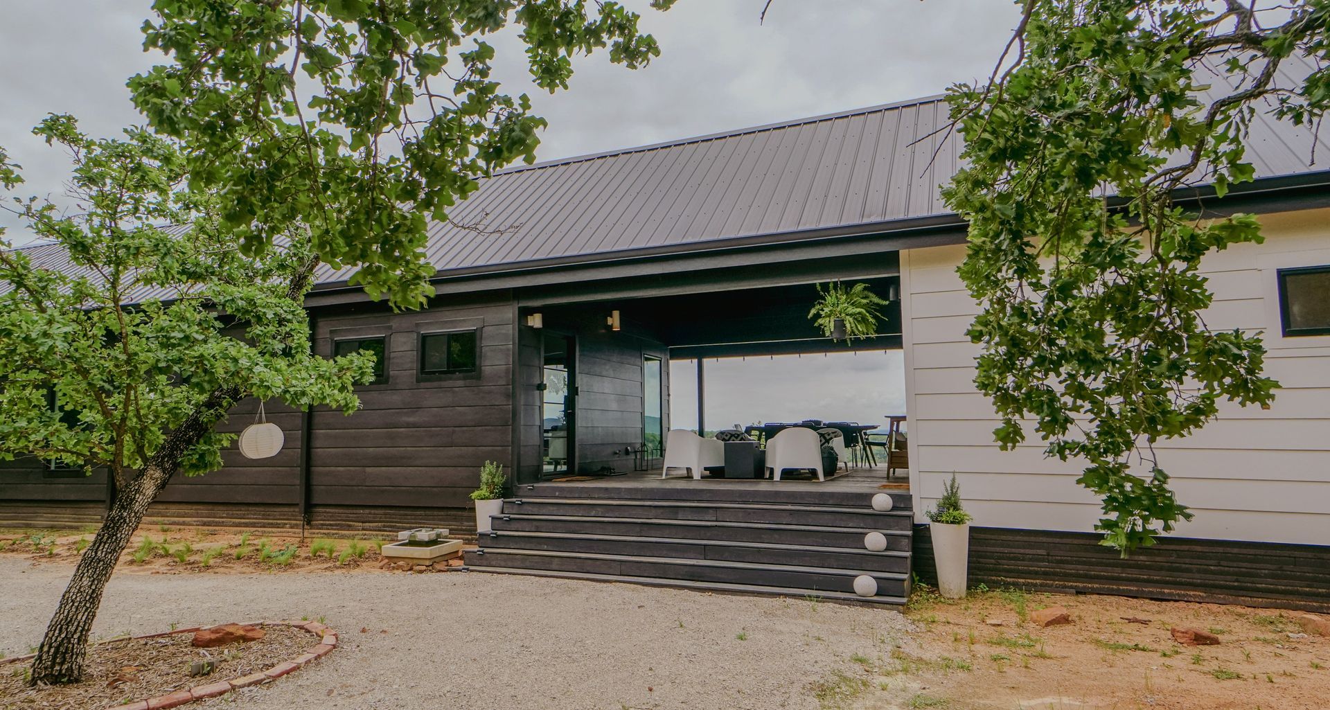 A modern house with a dark roof and a covered porch, set among trees on a cloudy day.