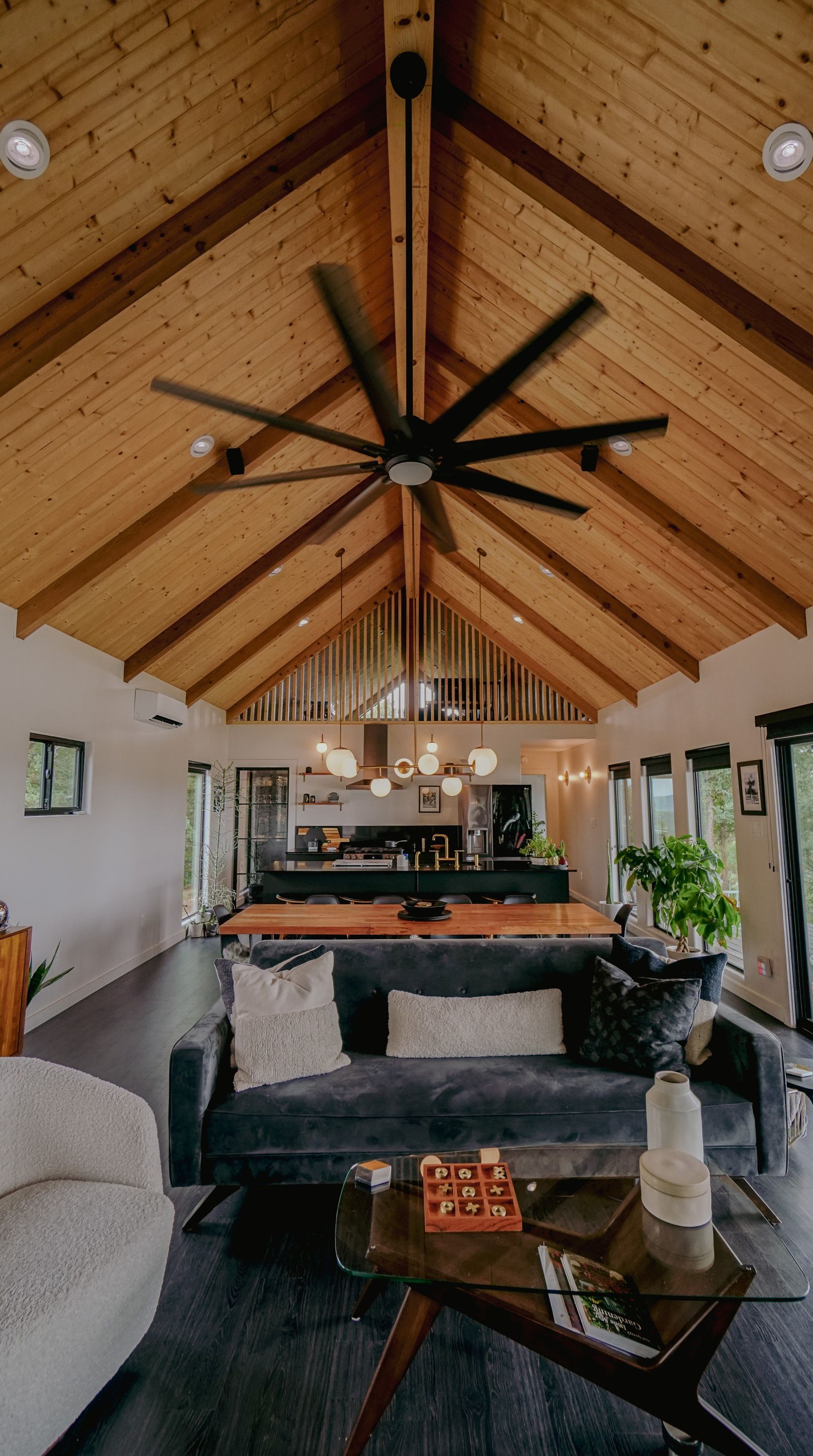 Living room with high wood-beamed ceiling. Large fan, dark couch, coffee table, and open kitchen visible.