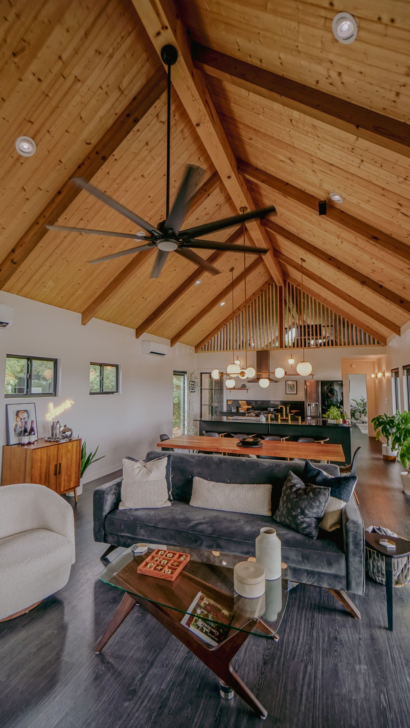 Living room with high wood-beamed ceiling, dark gray sofa, wooden coffee table, and open kitchen in the background.
