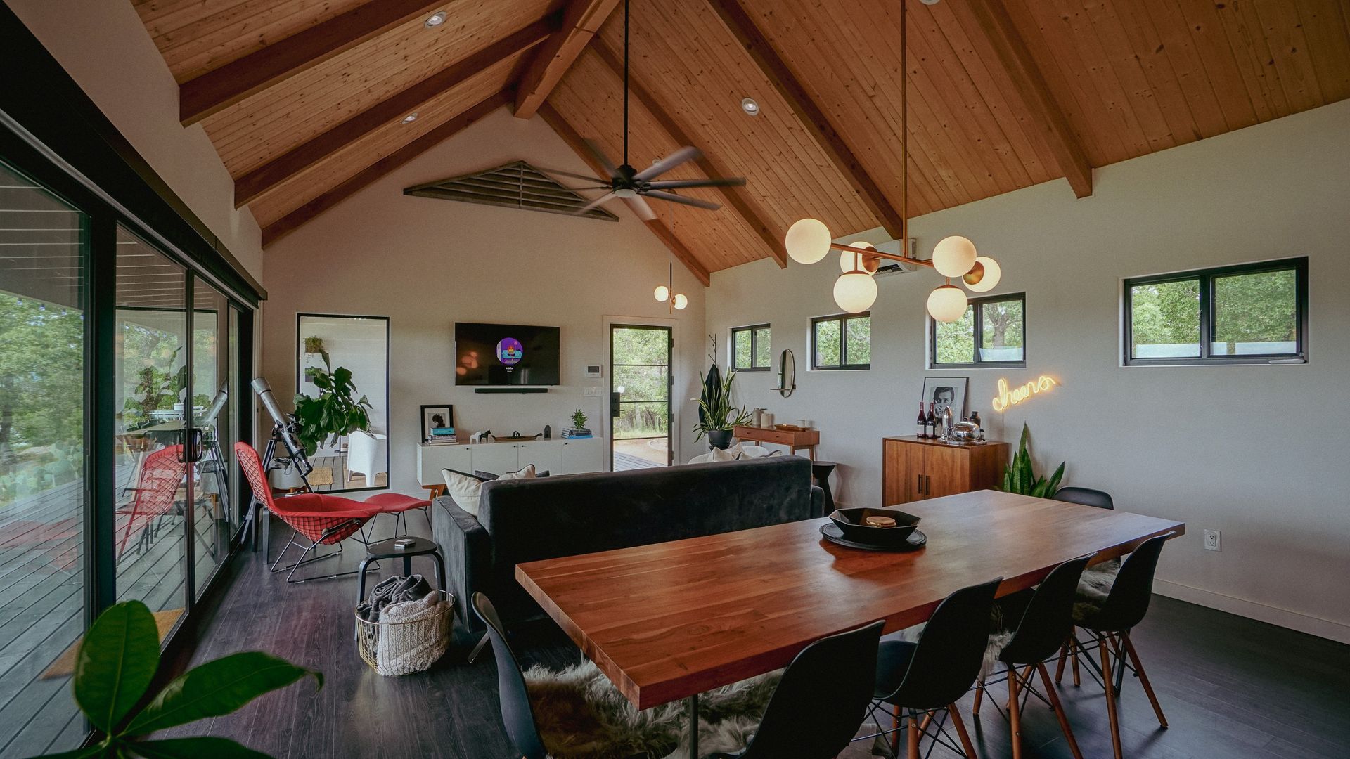 Modern open-concept living room with wood-paneled ceiling and large windows.  Dining table, couch, and wall-mounted TV.