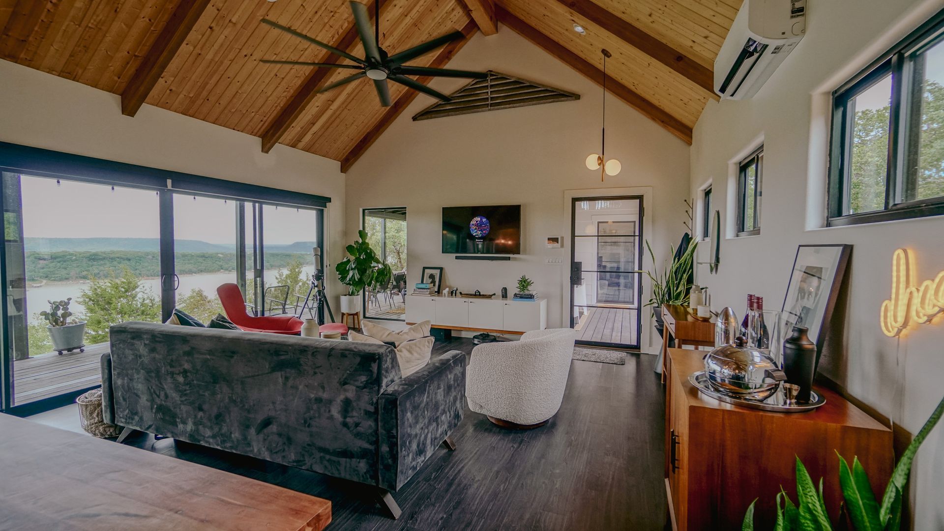 Living room with high wood ceiling, sliding glass doors overlooking a lake, dark wood floors, modern furniture, and a fan.