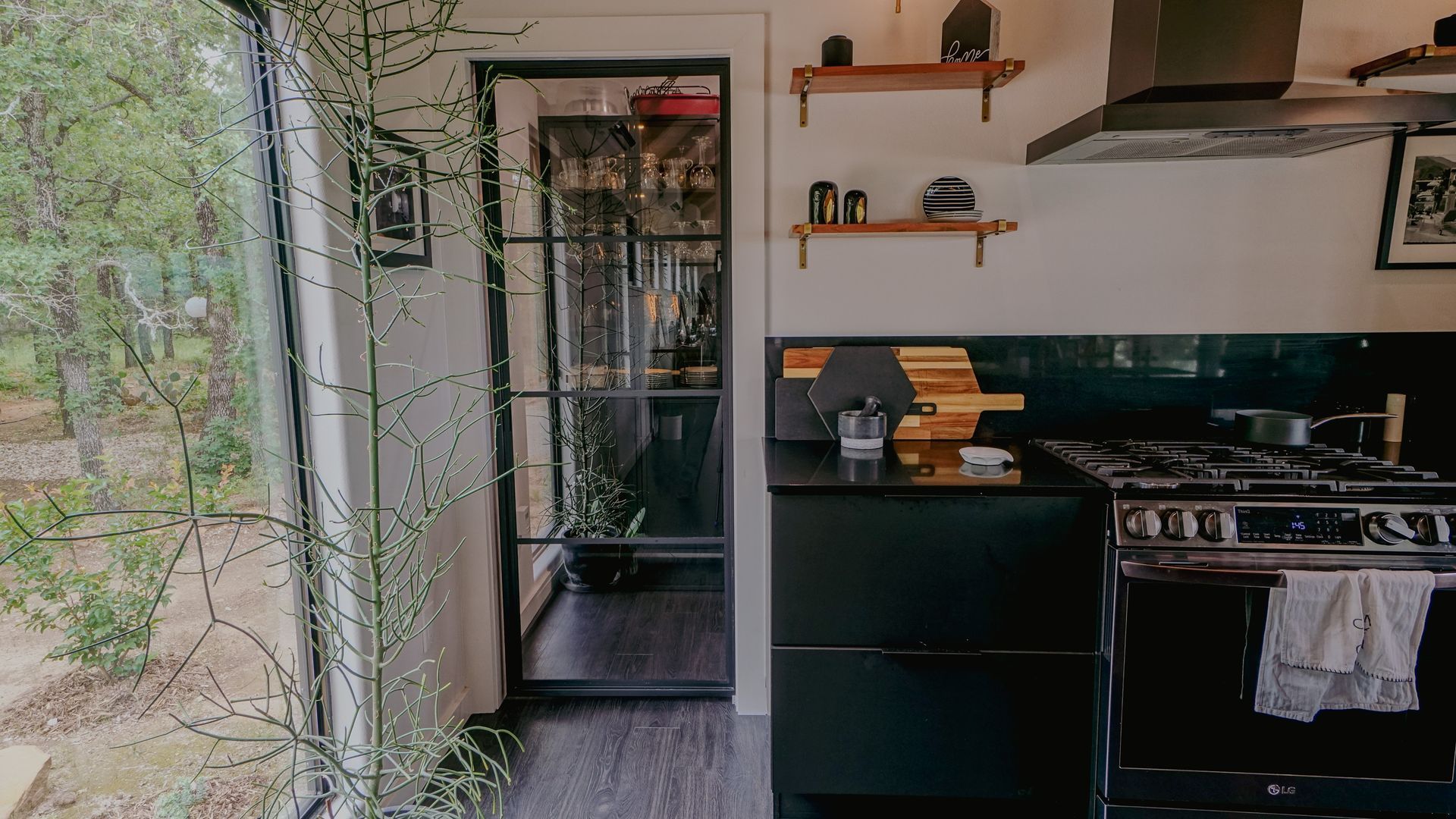 Modern kitchen with black cabinets and stove, glass door, shelves, and window looking out at trees.