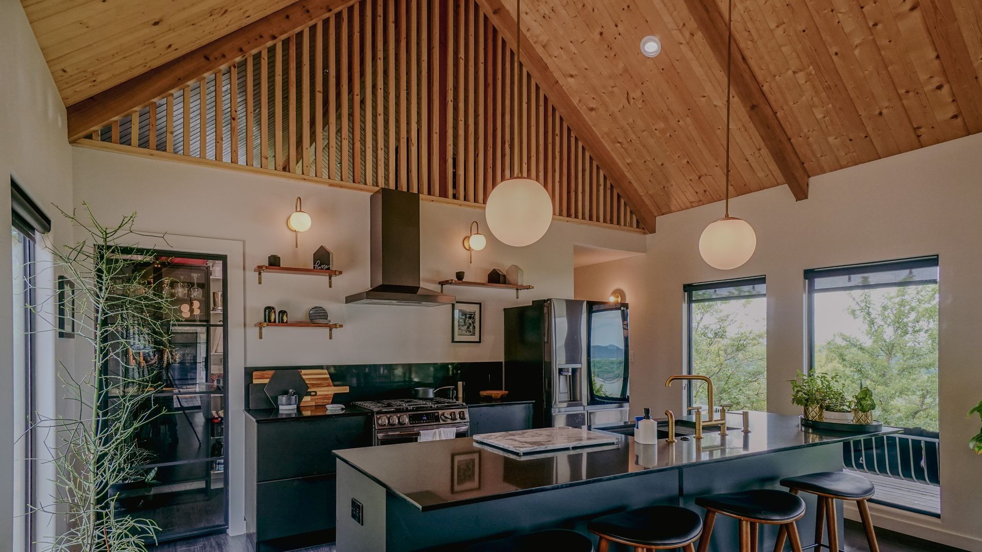 Modern kitchen with black island, wooden ceiling, and large windows with a view.