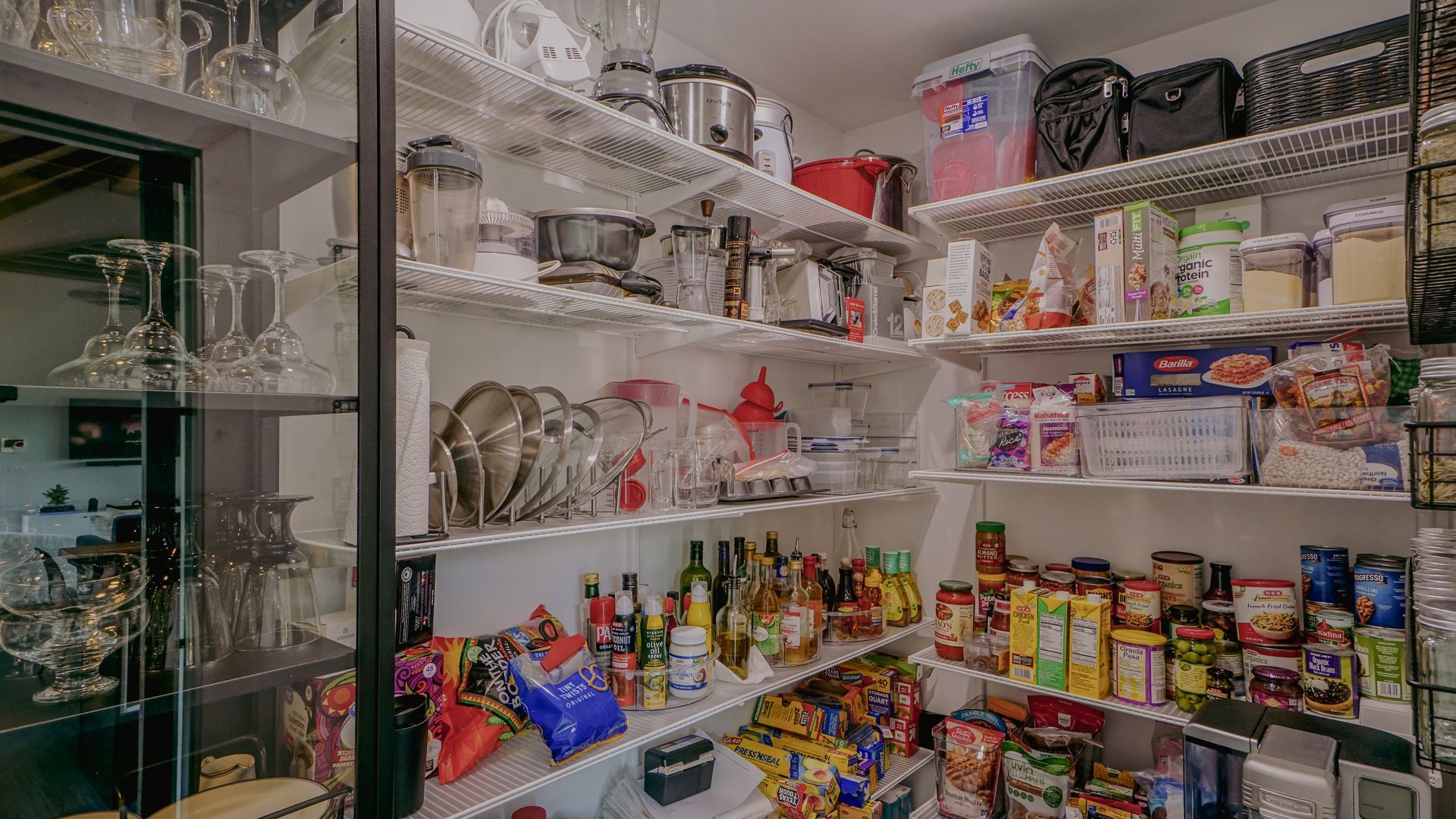 Pantry filled with food and kitchen appliances on white shelves. Glassware in cabinet on left.