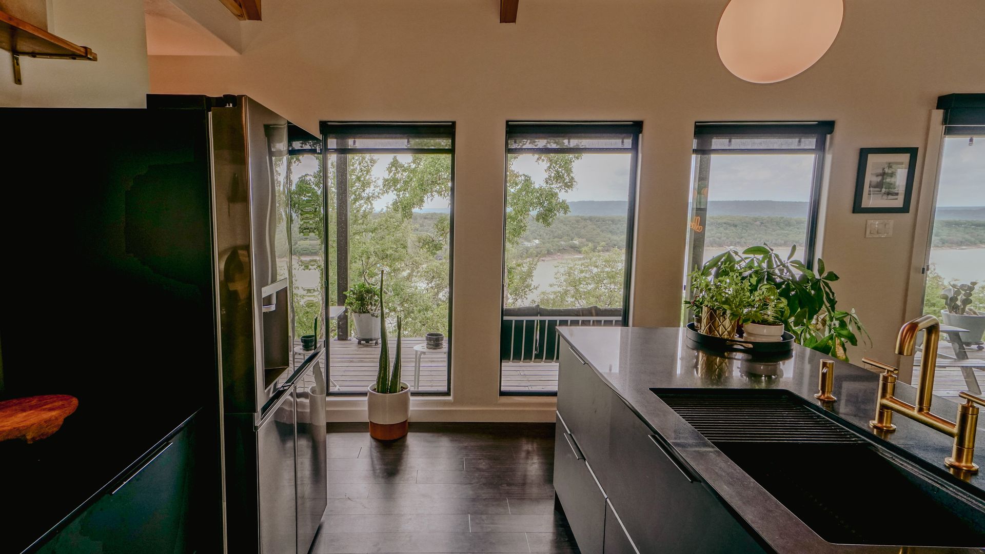 Modern kitchen with black cabinetry, stainless steel fridge, and windows overlooking greenery and water.
