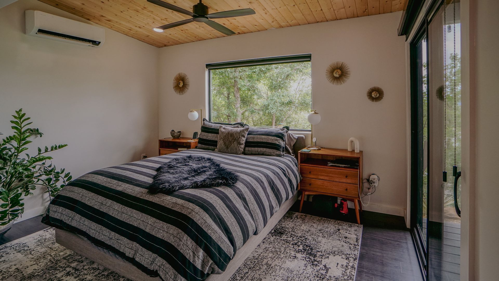Bedroom with a striped bed, wood-paneled ceiling, and large window overlooking greenery; two nightstands.