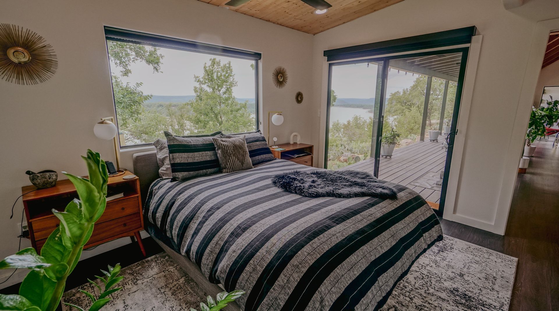 Bedroom with bed, large window, and sliding glass door to a porch overlooking a forest and lake.