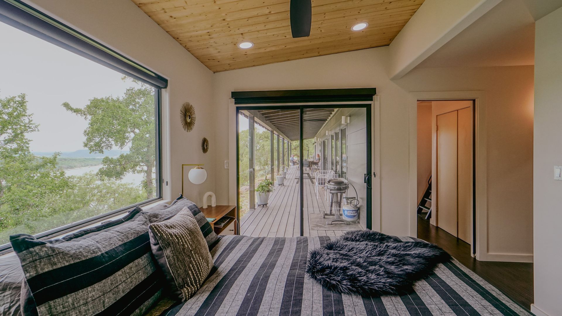 Bedroom with large windows overlooking a lake, sliding door to a deck, and a striped bed.