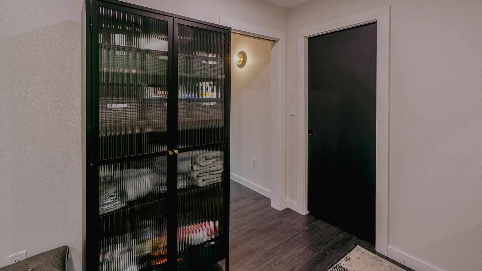 Black cabinet with glass doors in a hallway, dark door, hardwood floor, white walls.