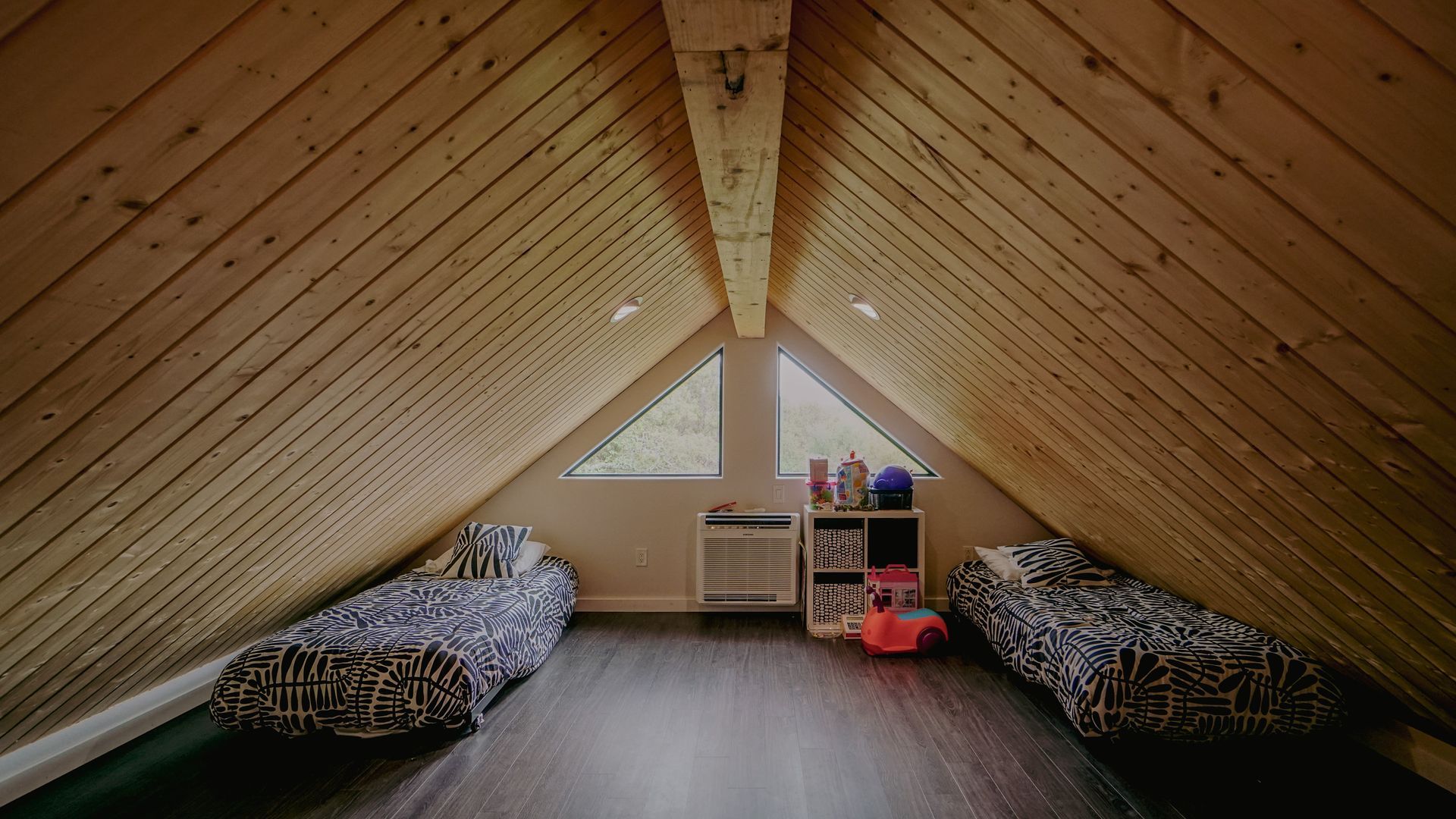 Attic bedroom with angled wooden ceiling, two twin beds, and triangular windows.