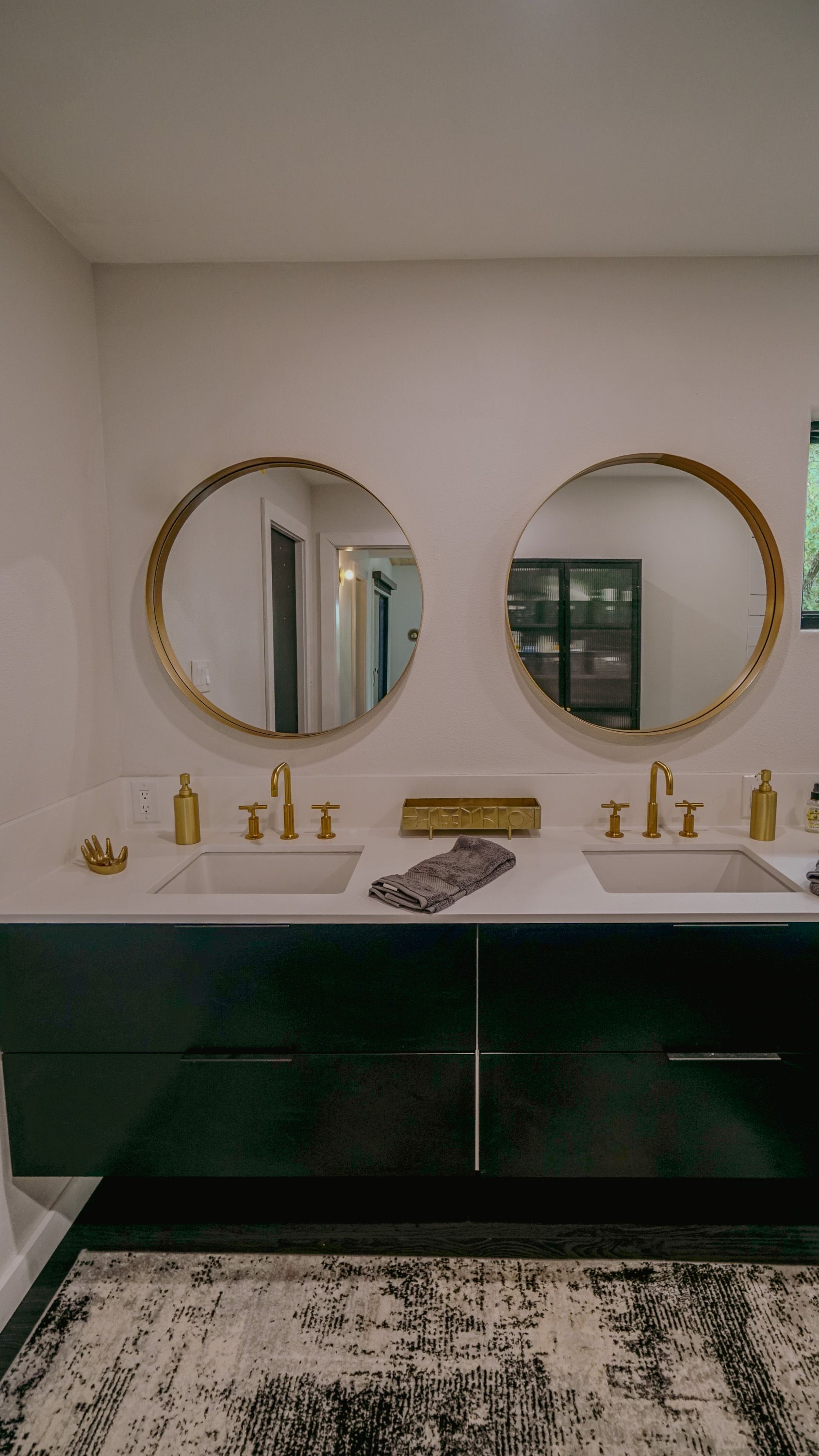 Modern bathroom with dual sinks, gold mirrors and fixtures, dark green vanity, and patterned rug.