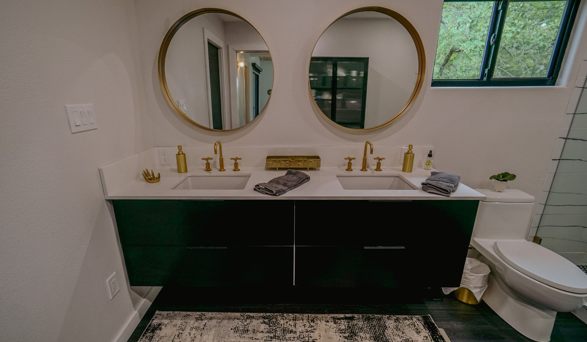 Bathroom with dark green vanity, two gold-framed mirrors, white countertop, gold fixtures, and a patterned rug.