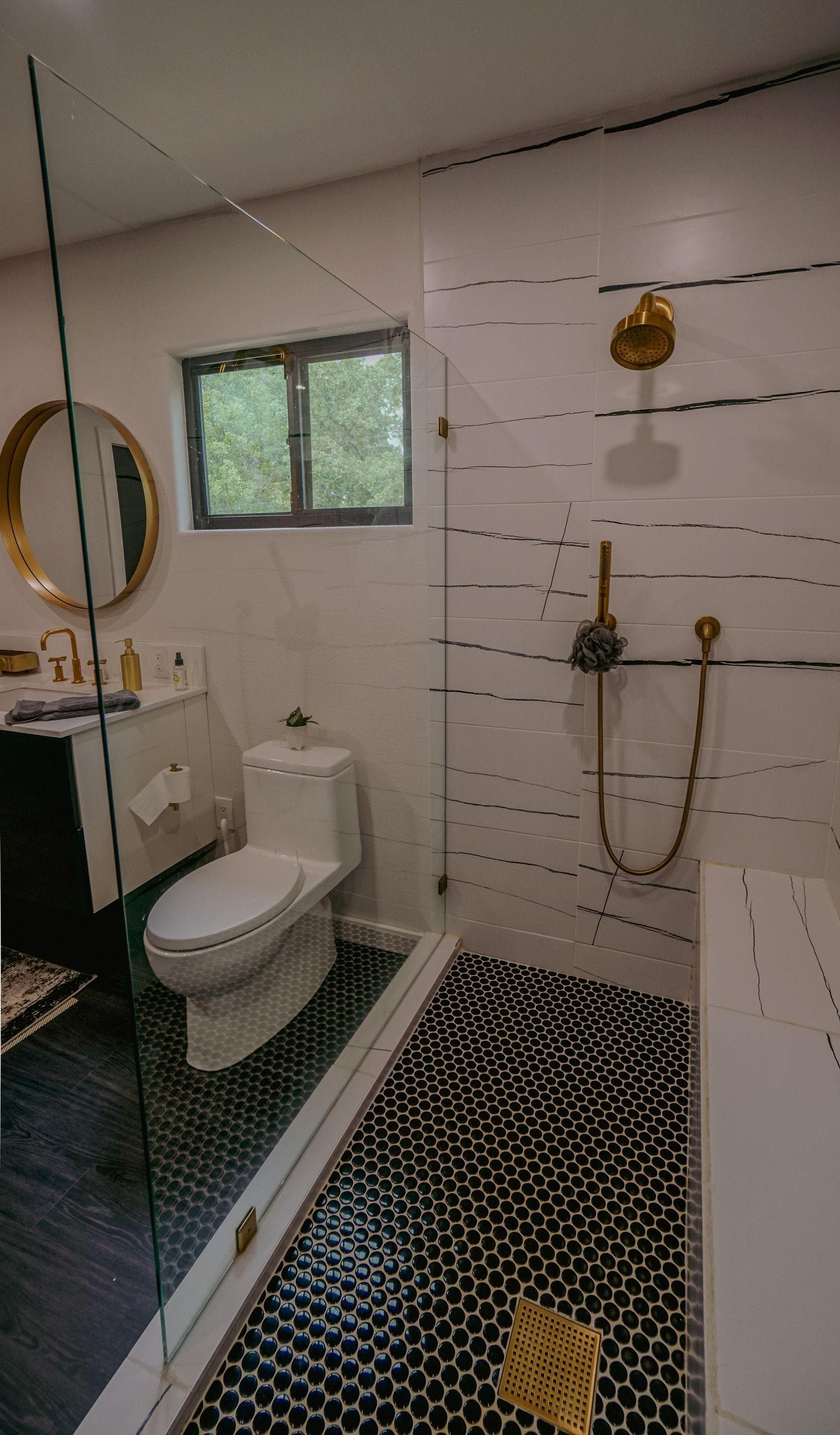 Modern bathroom with glass shower, black and white tile floor, gold fixtures, and a round mirror.