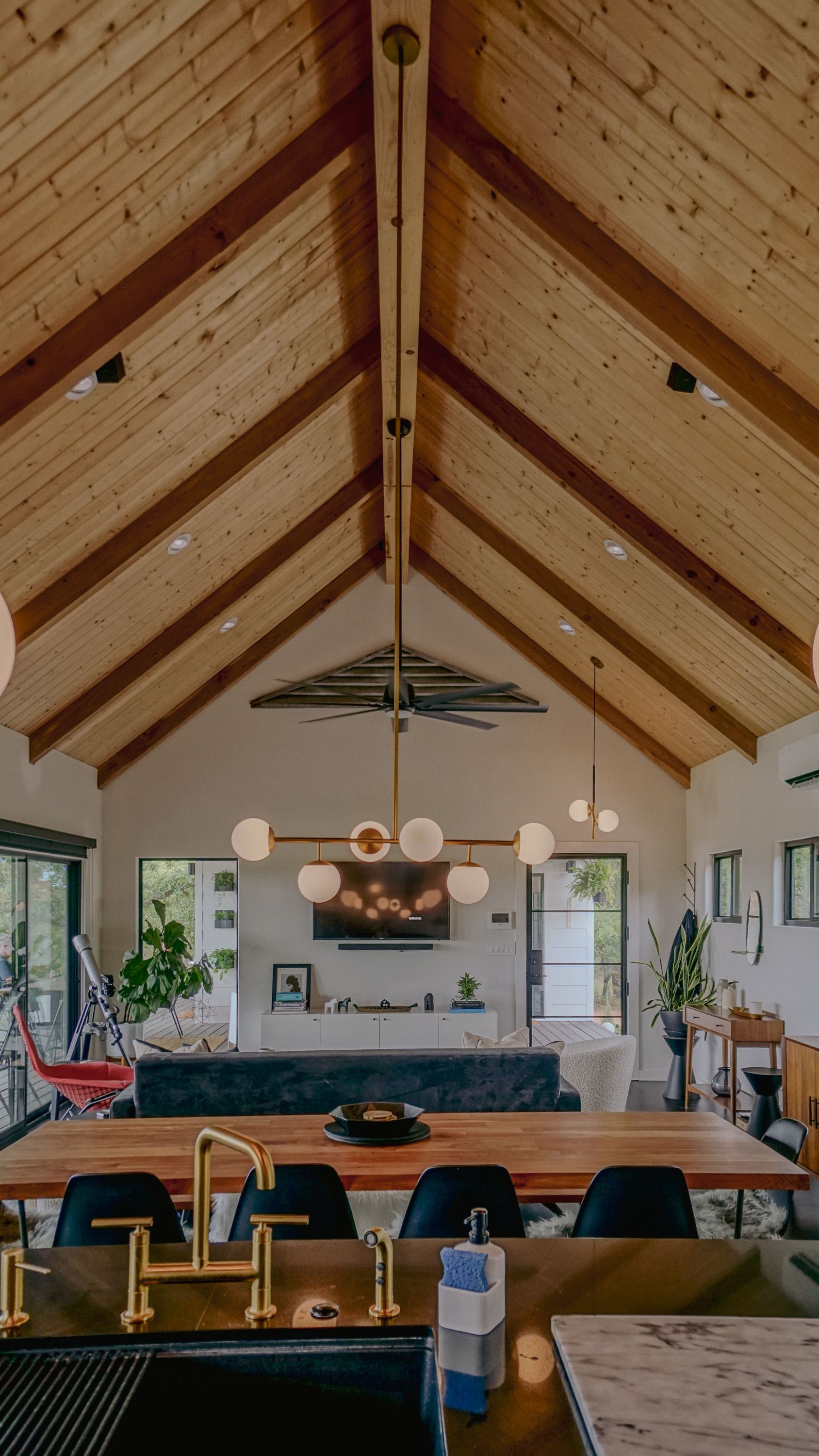 Living room with wood-paneled vaulted ceiling, chandelier, long table, fireplace, and large windows.