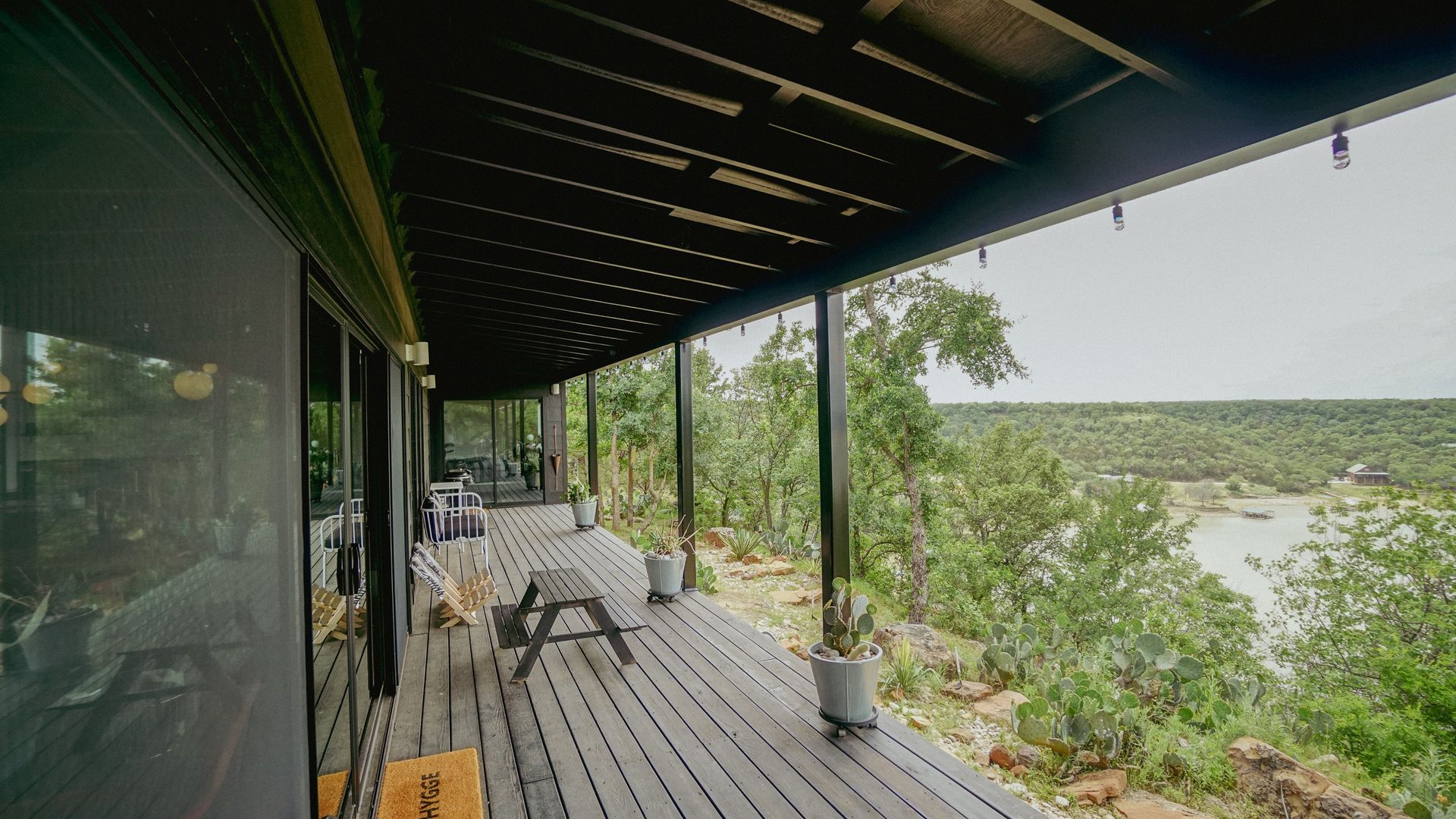 Covered wooden deck overlooking a tree-filled landscape. A-frame bench, potted plants visible.