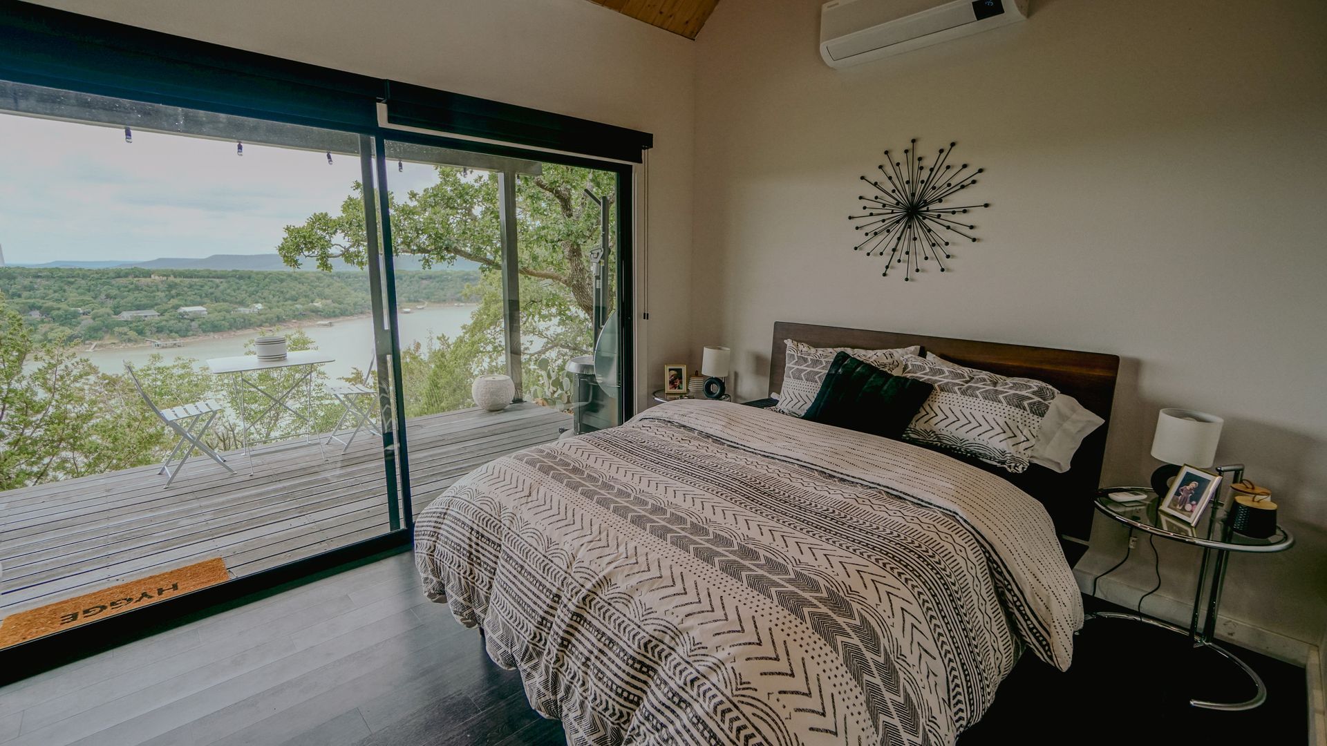 Bedroom with a large window overlooking a lake. Bed has patterned bedding, and there's a side table.