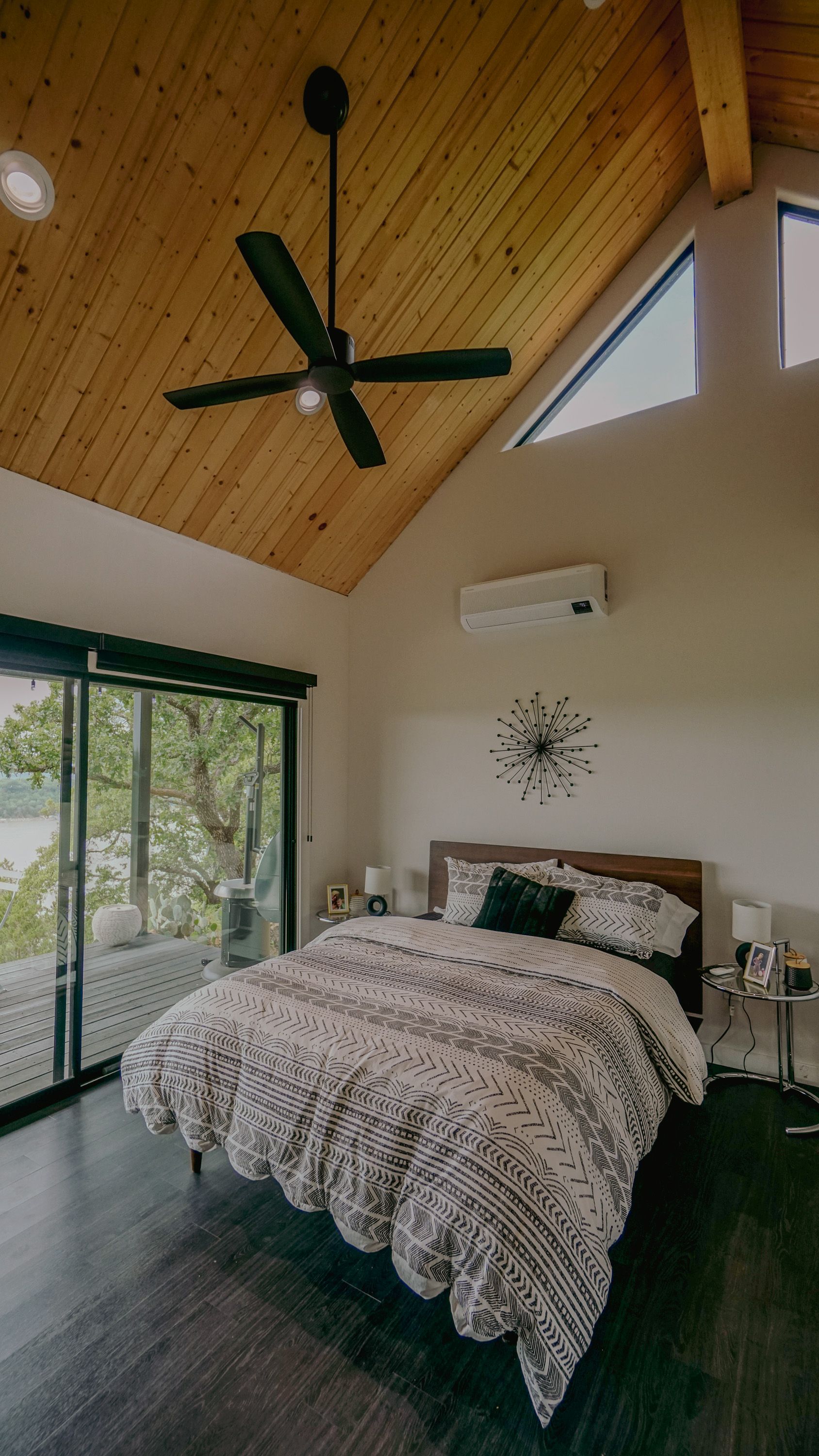 Bedroom with vaulted wooden ceiling, bed with patterned comforter, and glass door overlooking outdoor view.