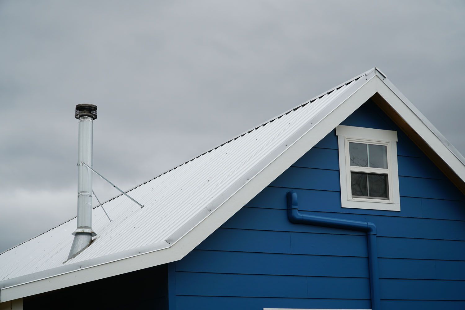 Blue house with white-framed window, metal roof, and chimney against a cloudy sky.