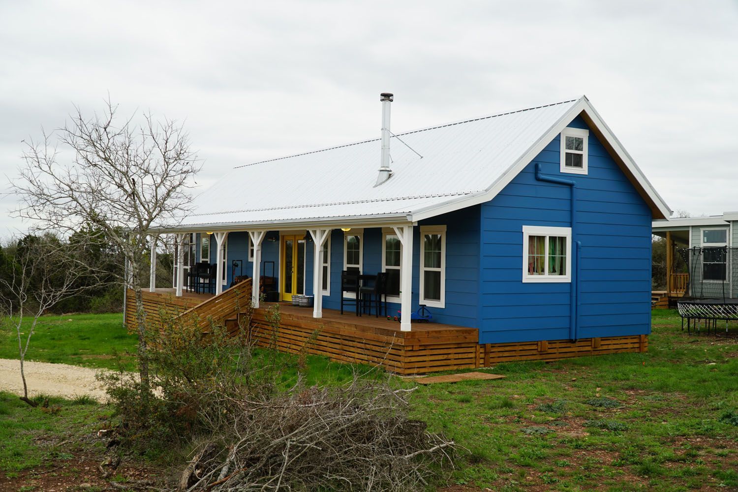 Blue cottage with a white porch and roof, surrounded by grass and a few trees.