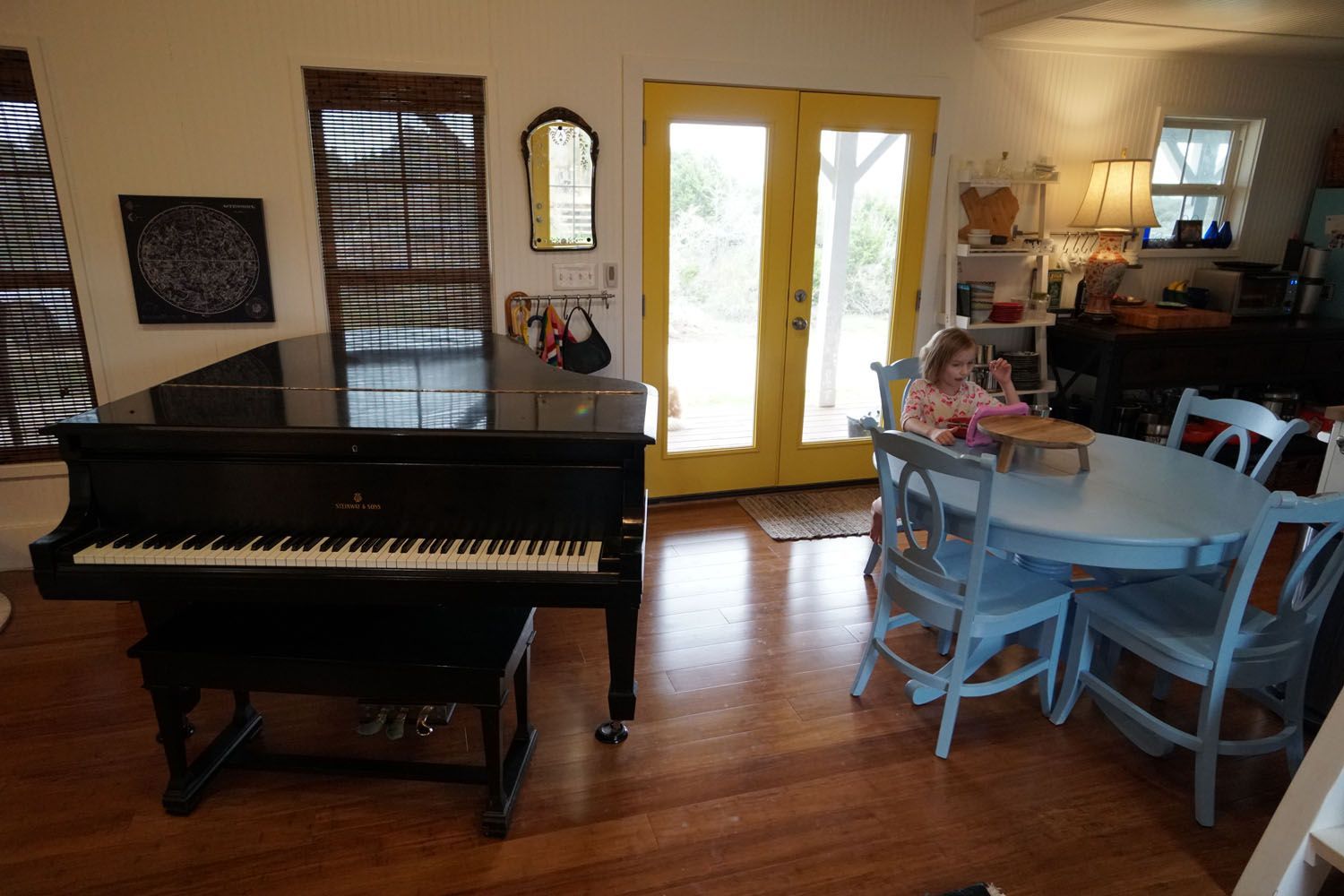A black piano sits in a room with a child at a blue table near yellow doors.