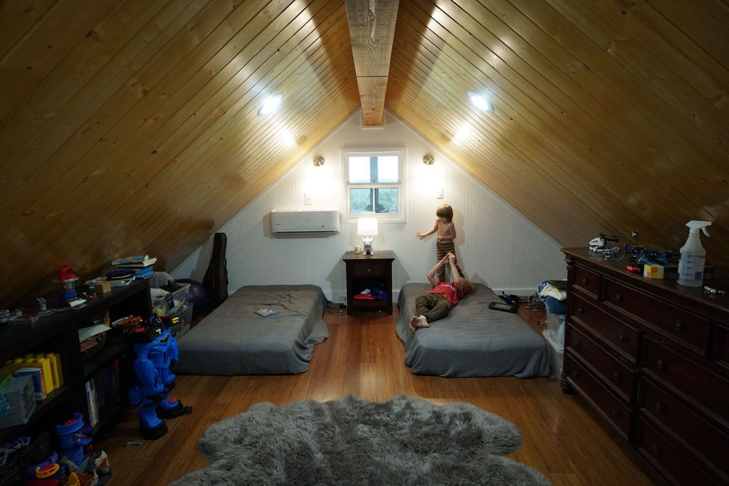 Two children playing in a wood-paneled attic bedroom with two beds, a rug, and a window.