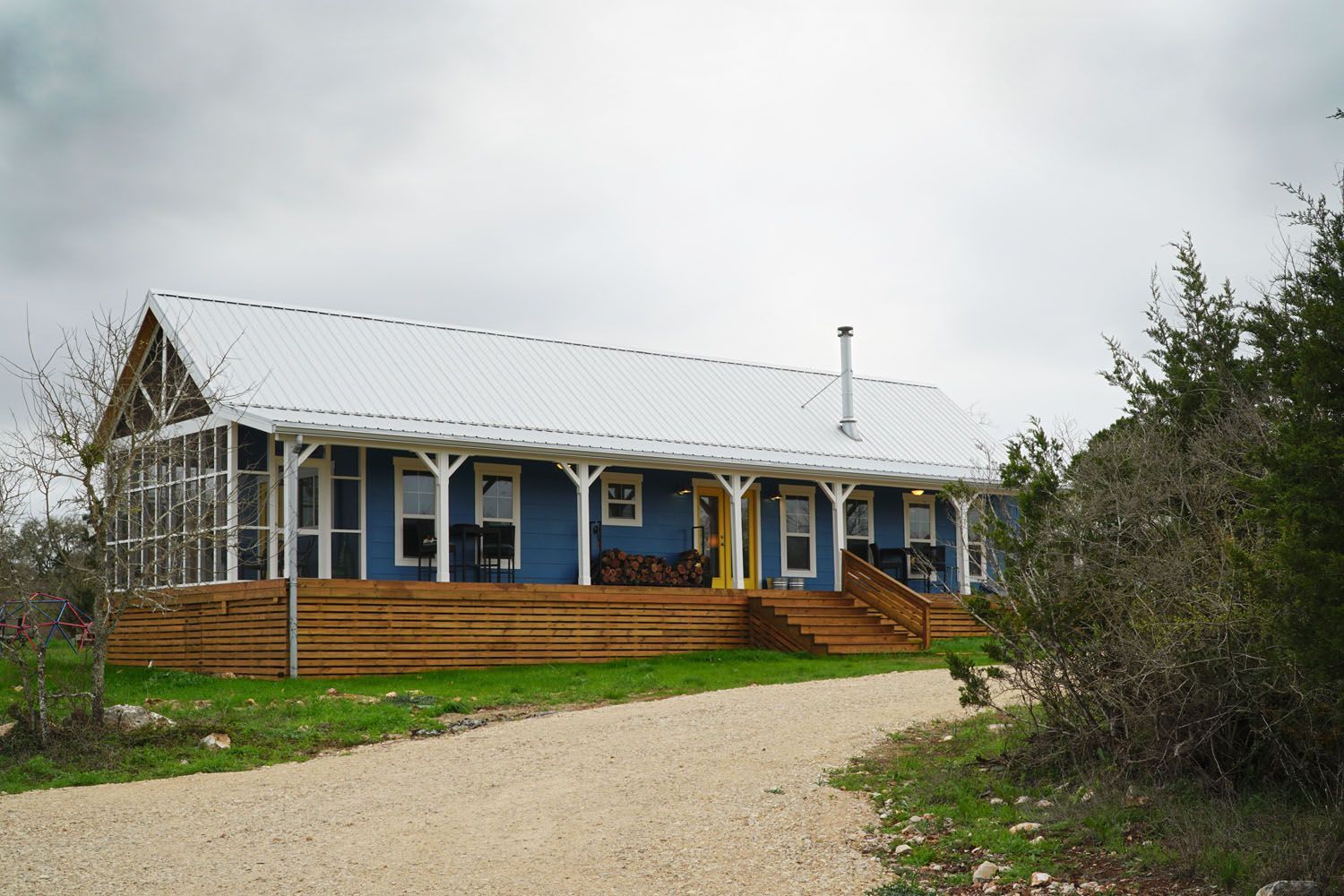 Blue house with porch and white metal roof, on a gravel driveway, surrounded by trees.