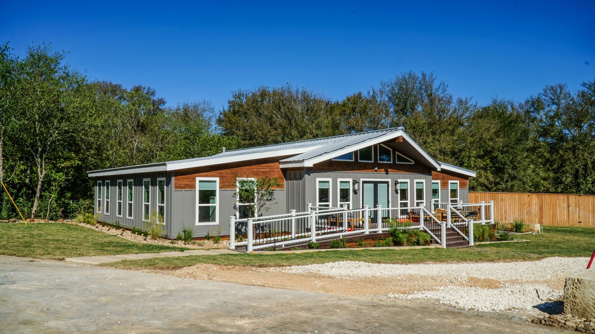 Single-story building with a ramp, white railing, and wooden accents; gray exterior and metal roof on a sunny day.
