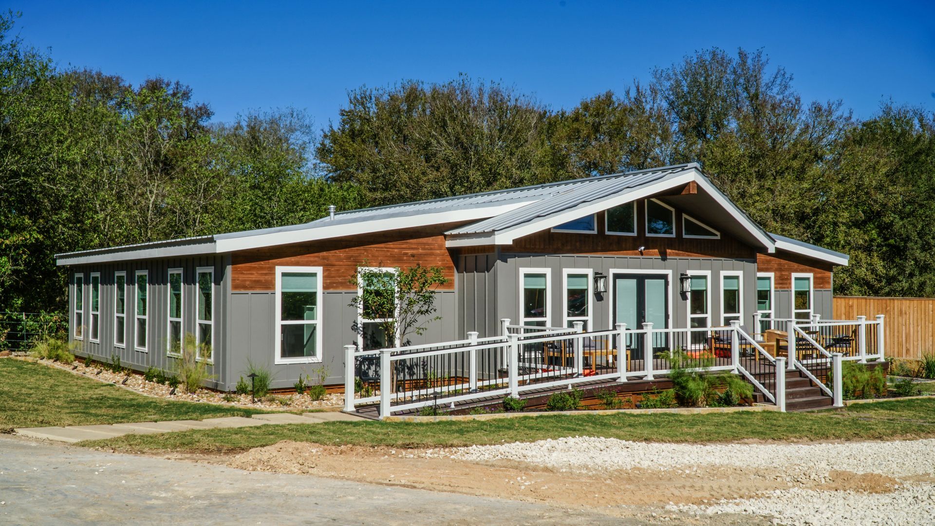 Gray house with a brown trim, white windows, and a wheelchair ramp, surrounded by trees under a clear sky.