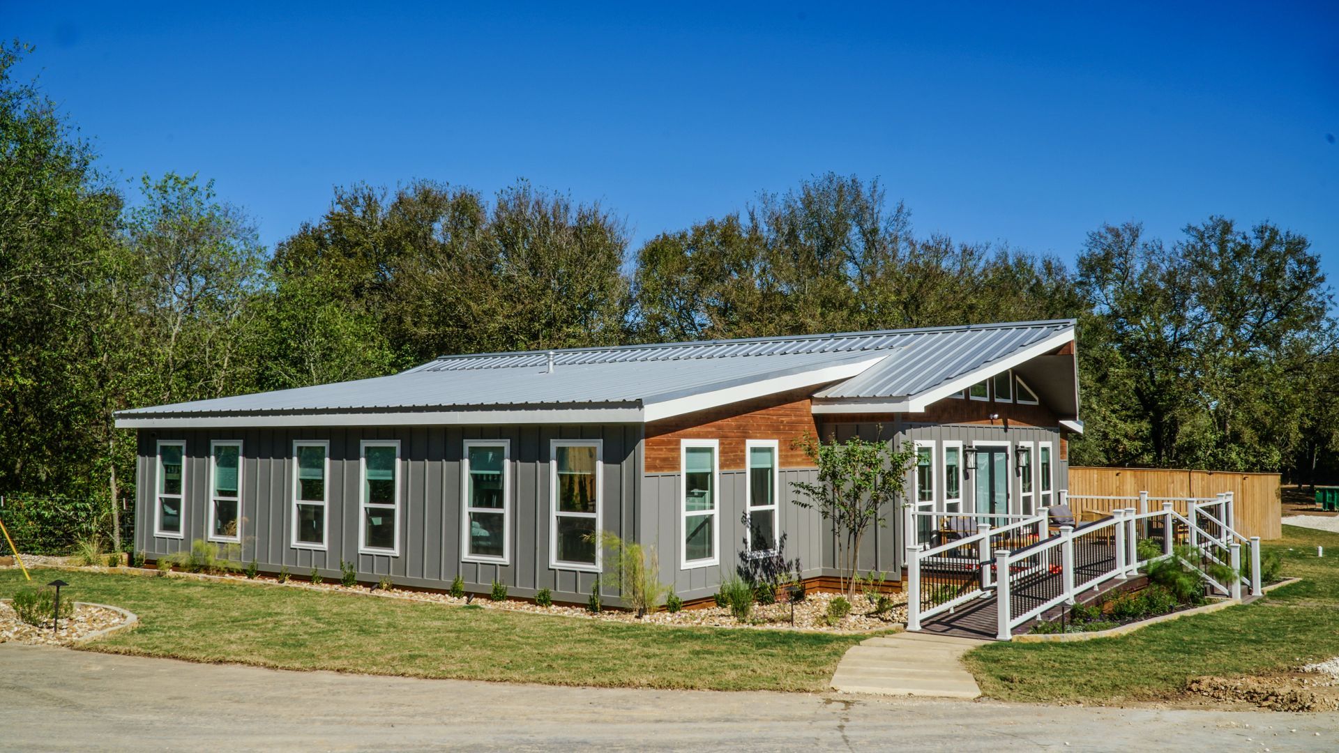 Grey building with angled metal roof, multiple windows, and wooden accents; ramp and walkway leading to the entrance.