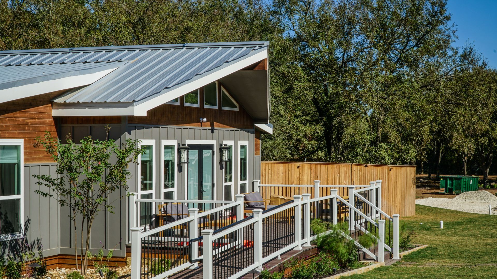 Modern cabin with gray siding, metal roof, and white porch railing; green trees in the background.