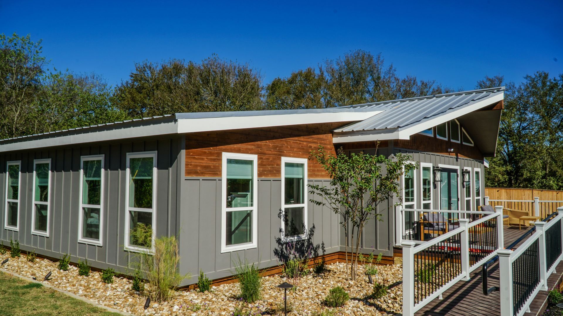 Modern gray and brown house with a sloping roof, large windows, and a deck in a sunny outdoor setting.