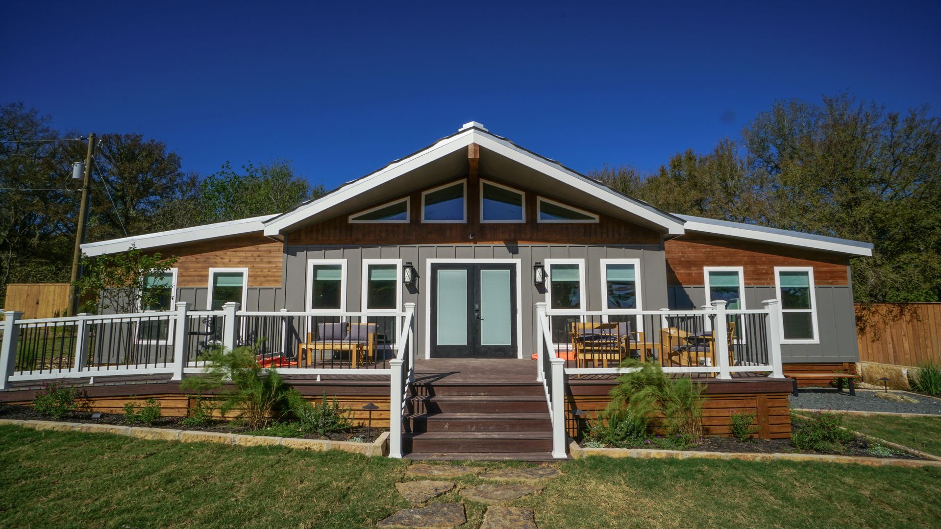 Ranch-style house with gray siding, white trim, and a wooden deck. The front door is flanked by windows, under a blue sky.