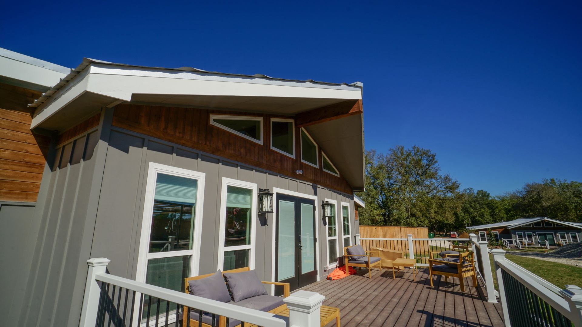 Gray house with a deck. The house has a white-trimmed roof with brown wood accents and several windows. Blue sky overhead.