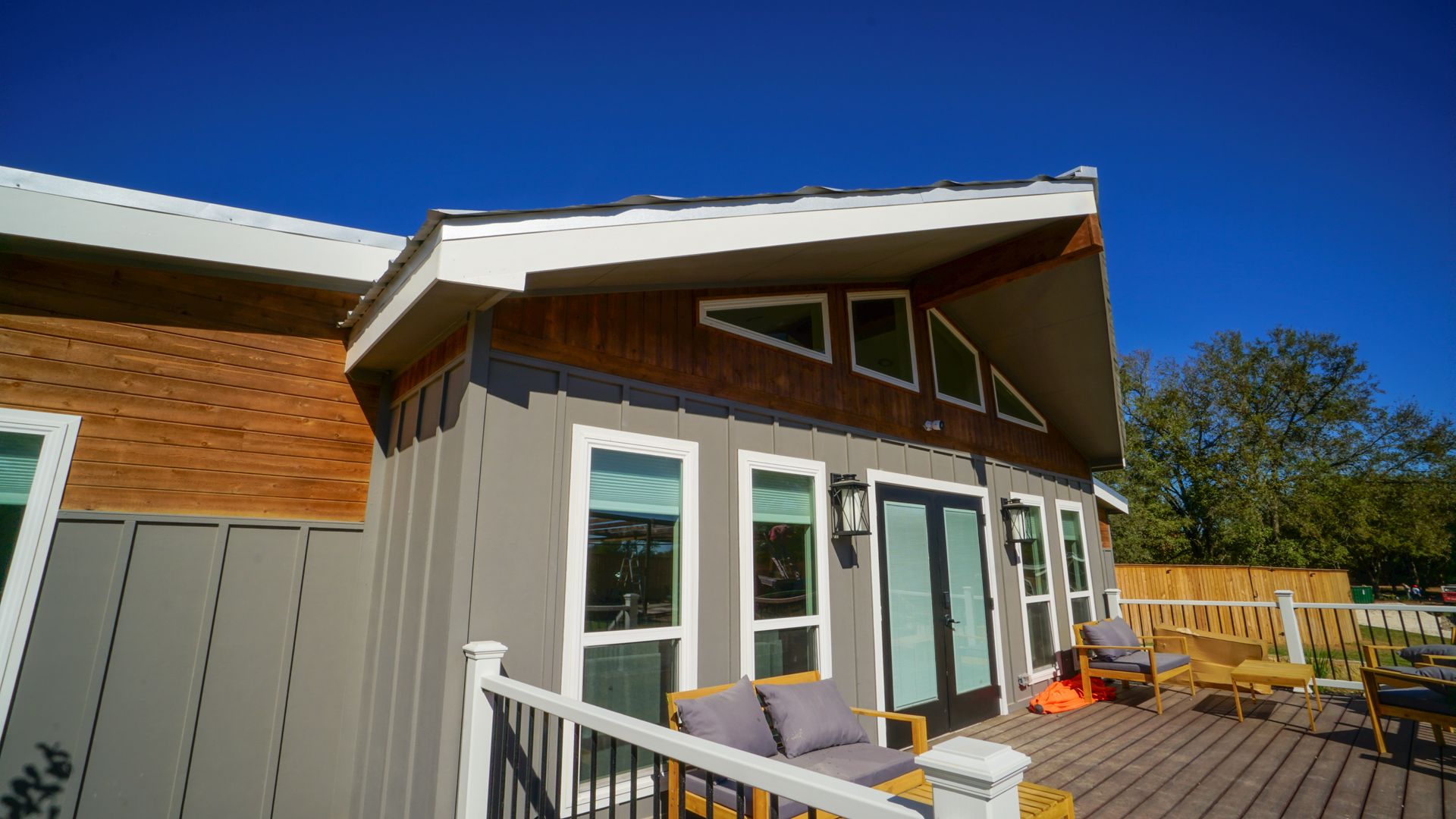 Modern gray home with wooden accents and windows, sunny outdoor deck with furniture.
