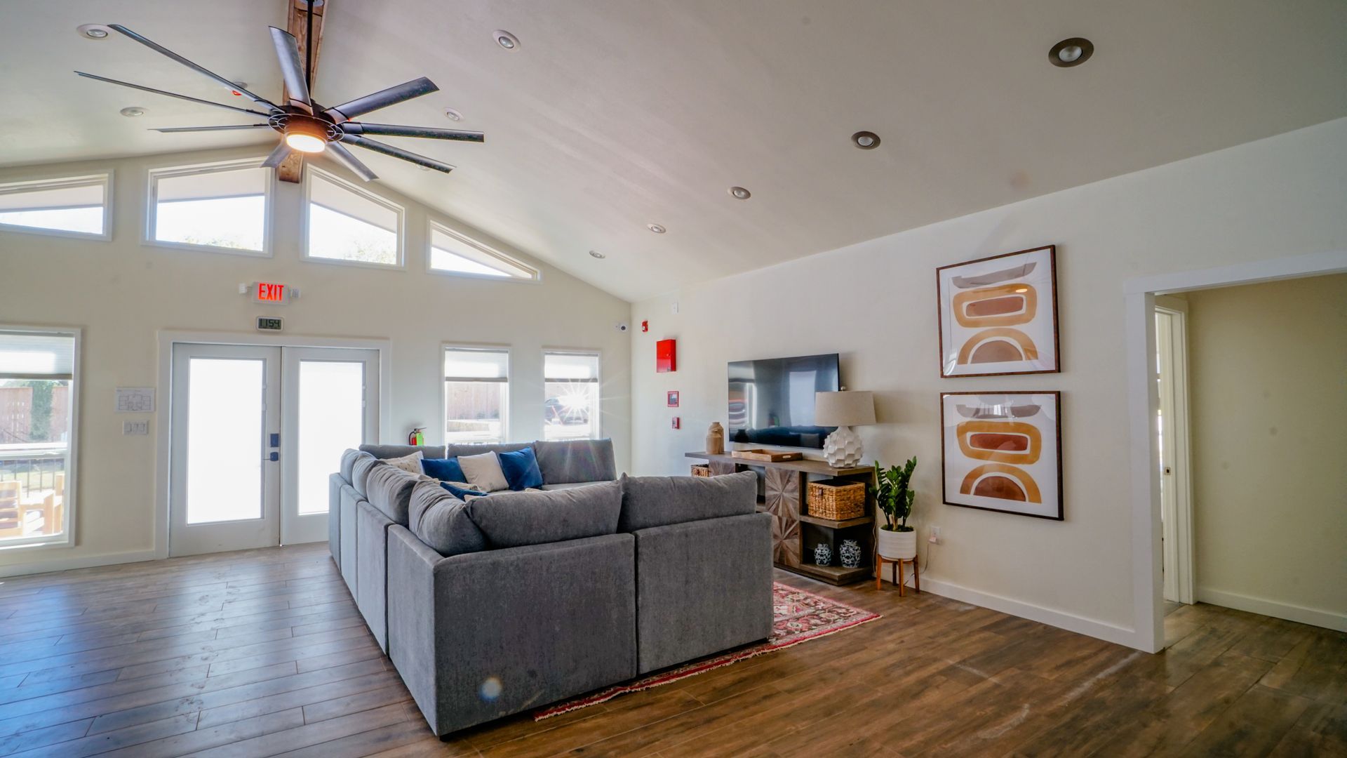 Spacious living room with gray sectional sofa, TV, and large windows. Brown hardwood floors and high ceiling.