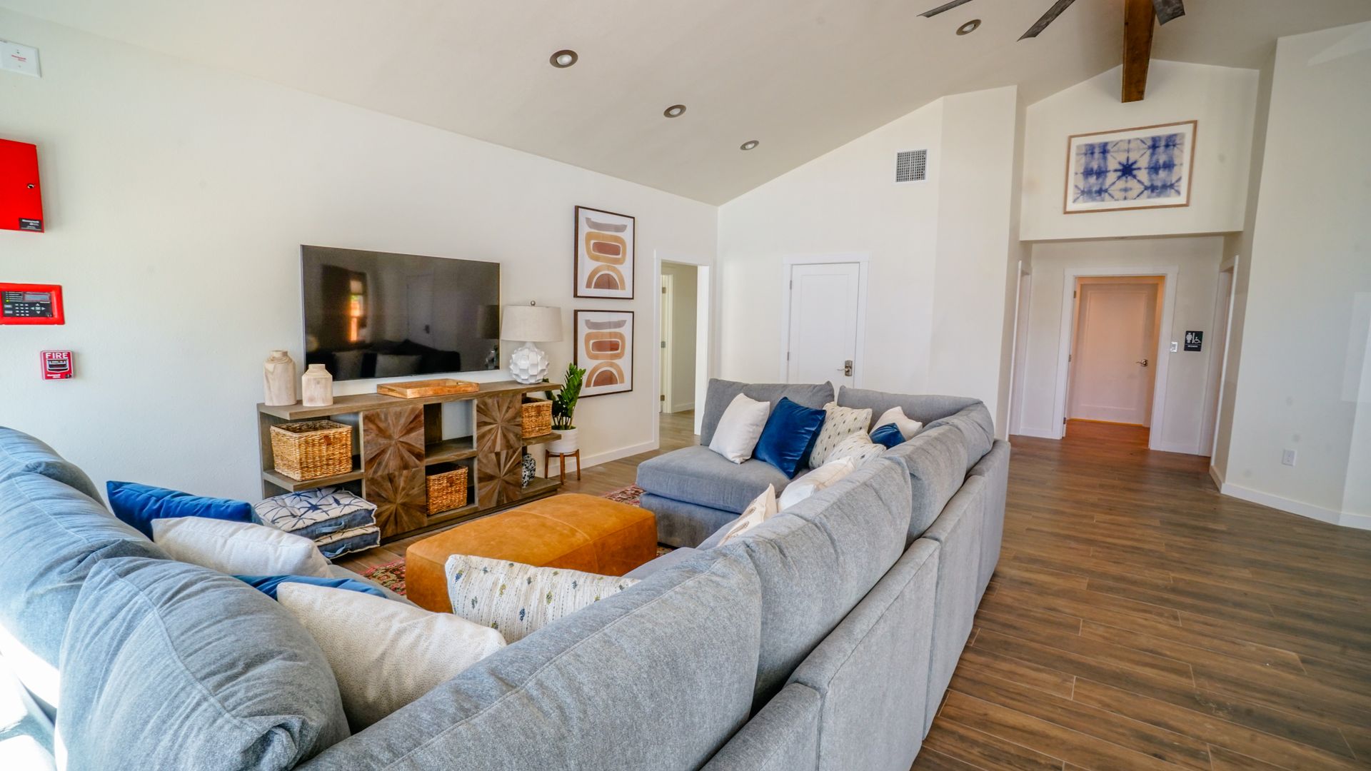 Spacious living room with a gray sectional, orange ottoman, wood entertainment center, and white walls.