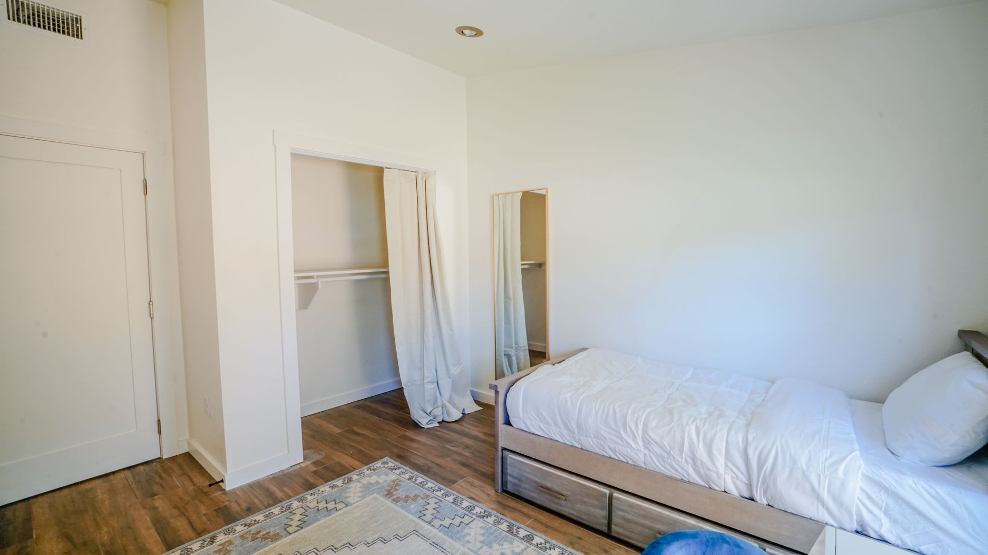 Bedroom interior with white walls, a bed, closet, and rug on a hardwood floor.