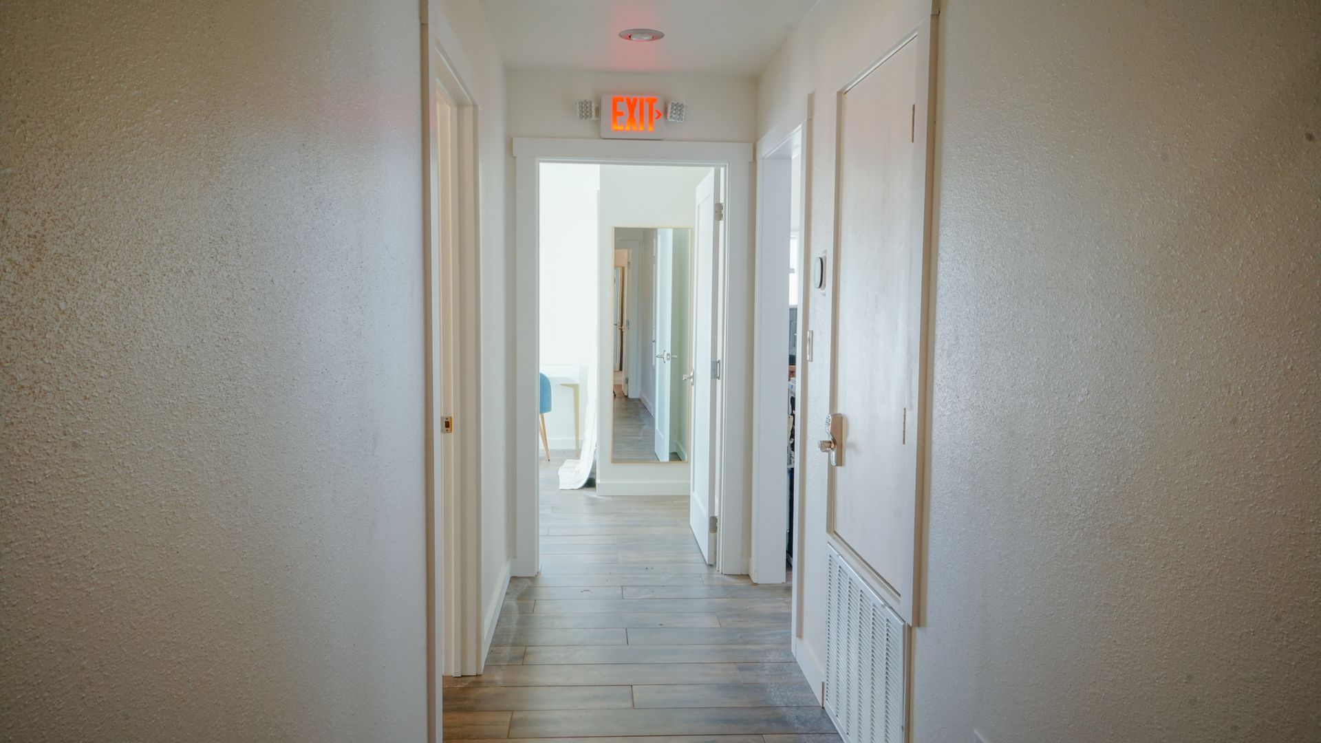 Hallway with light-colored walls and wooden floor. Open doors lead to rooms; exit sign above a doorway.