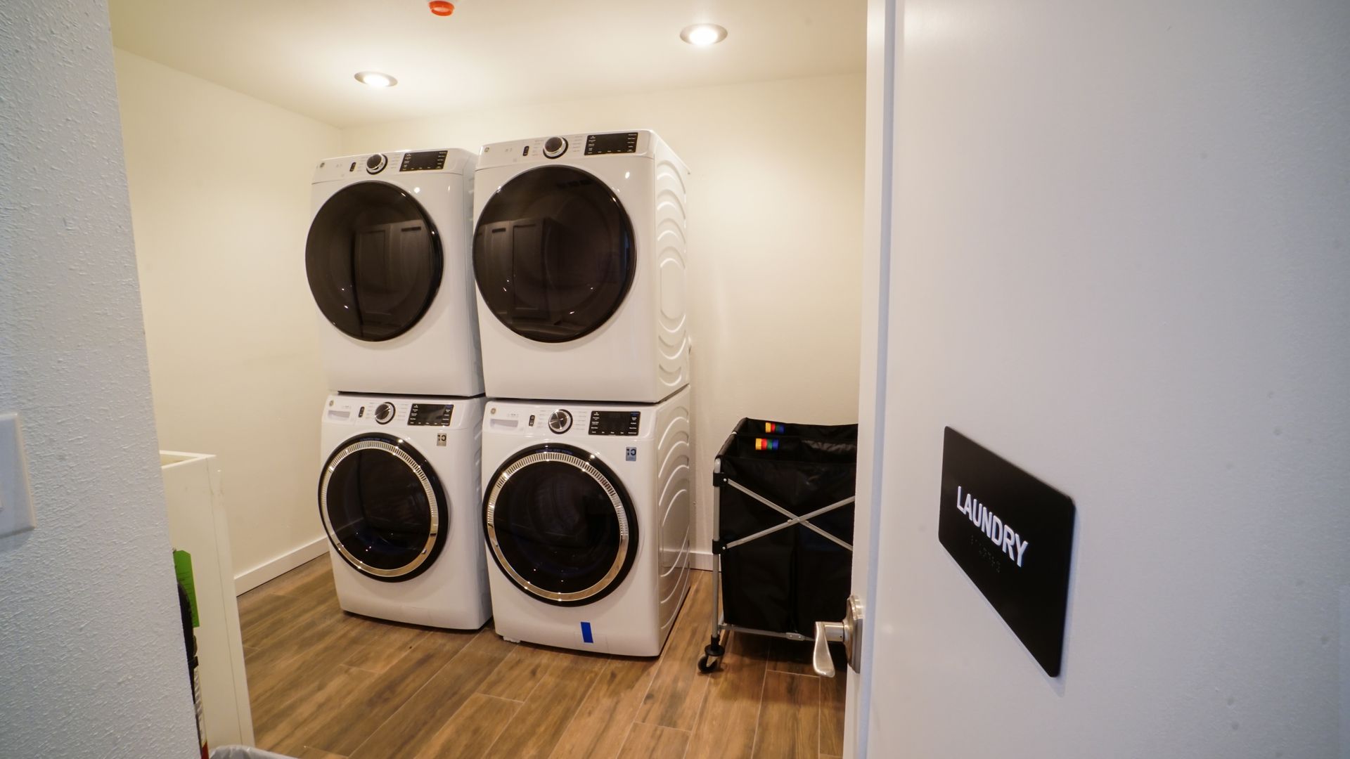 Laundry room with stacked white washers and dryers, black laundry cart, and open door.