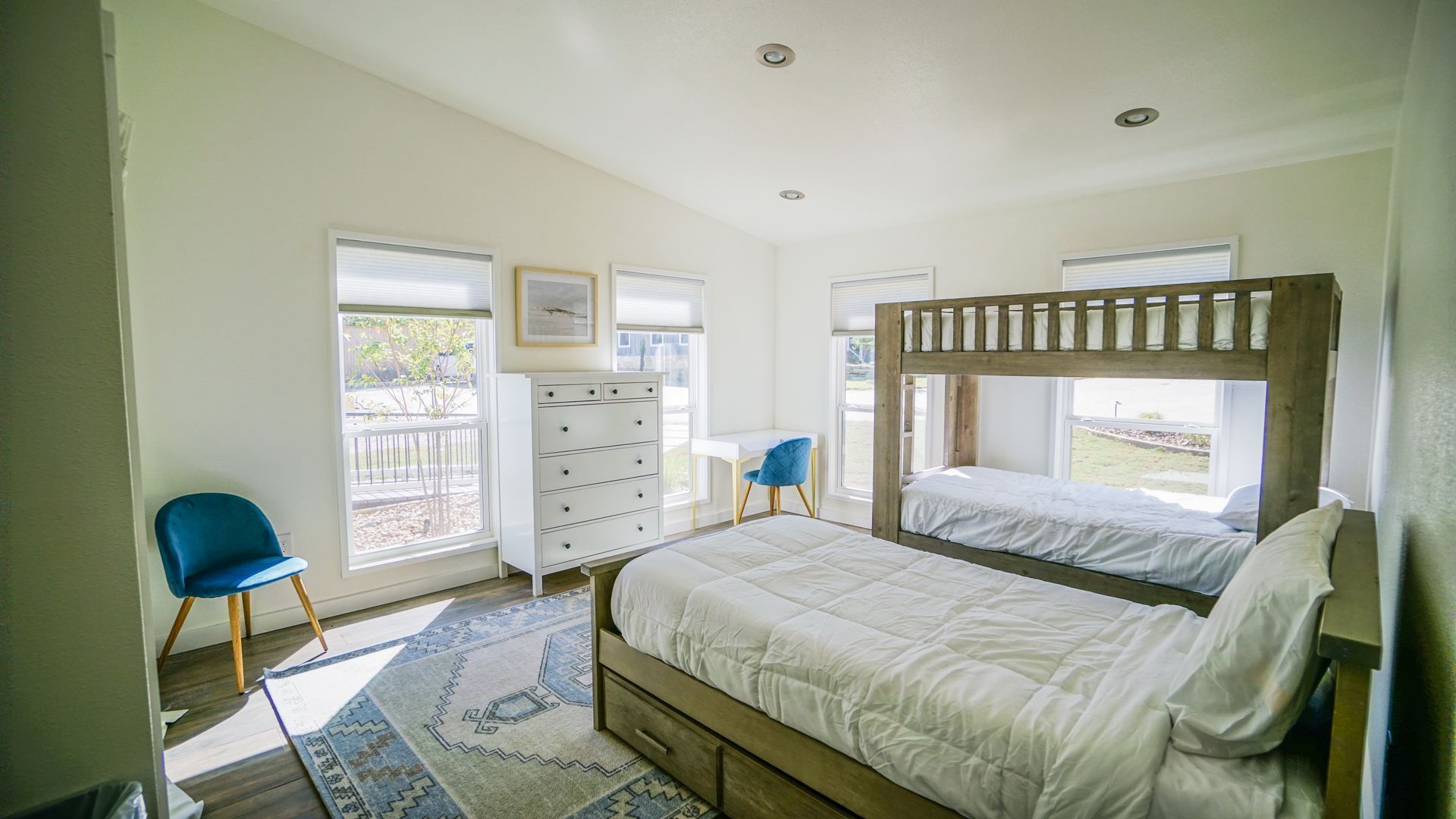 Bedroom with bunk bed, twin bed, white dresser, and blue chairs. Bright room with large windows.