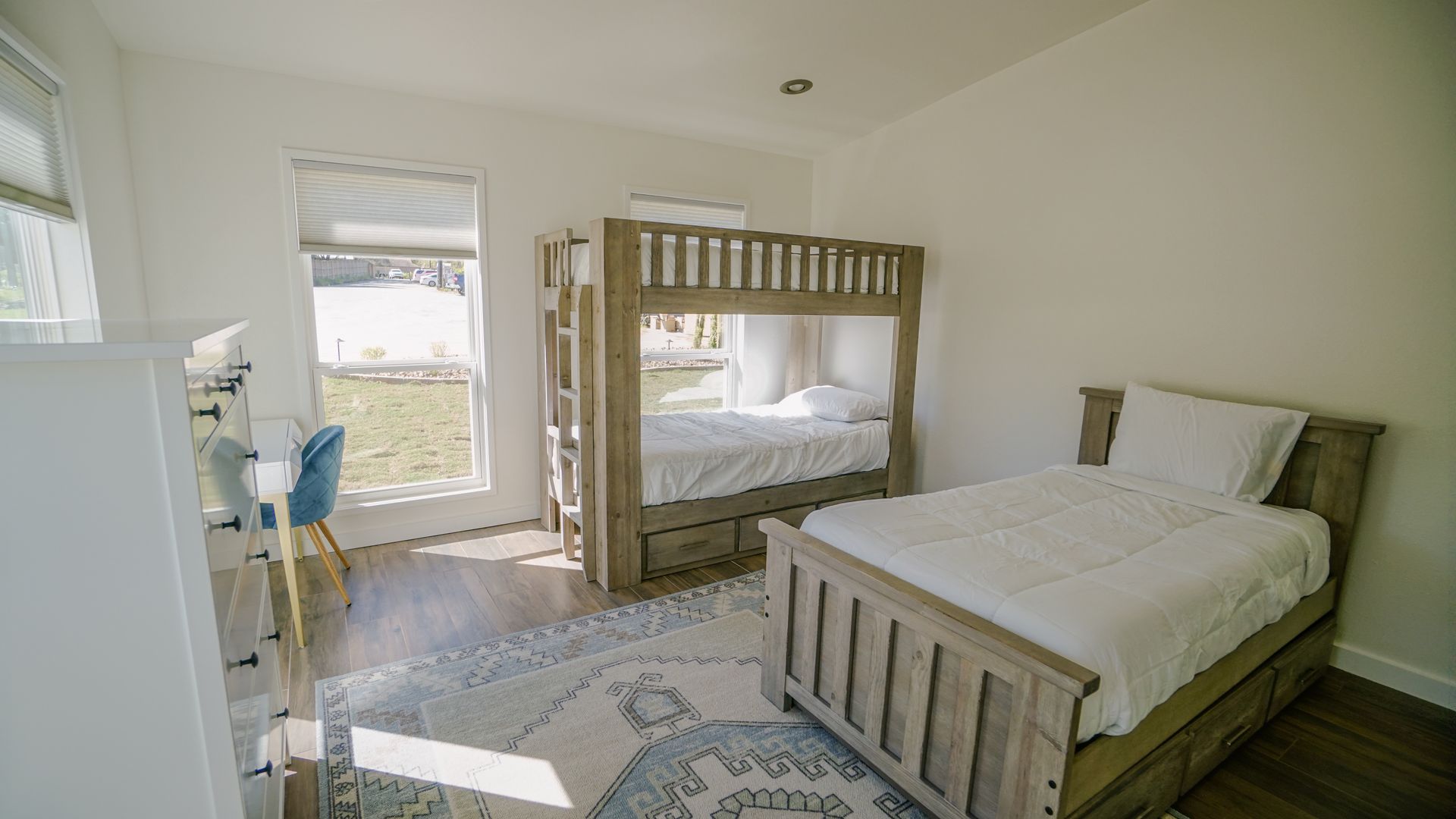 Bedroom with bunk beds, a single bed, desk, and area rug. White walls, wood furniture, window with blinds.