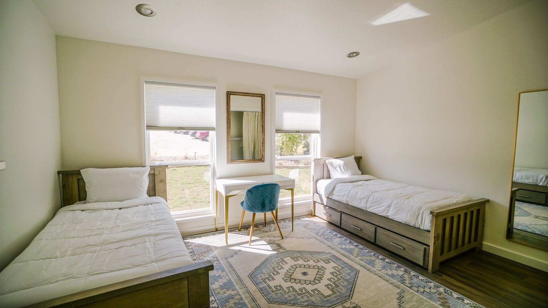 Bedroom with two twin beds, desk, rug, windows, and mirror. White and beige decor.