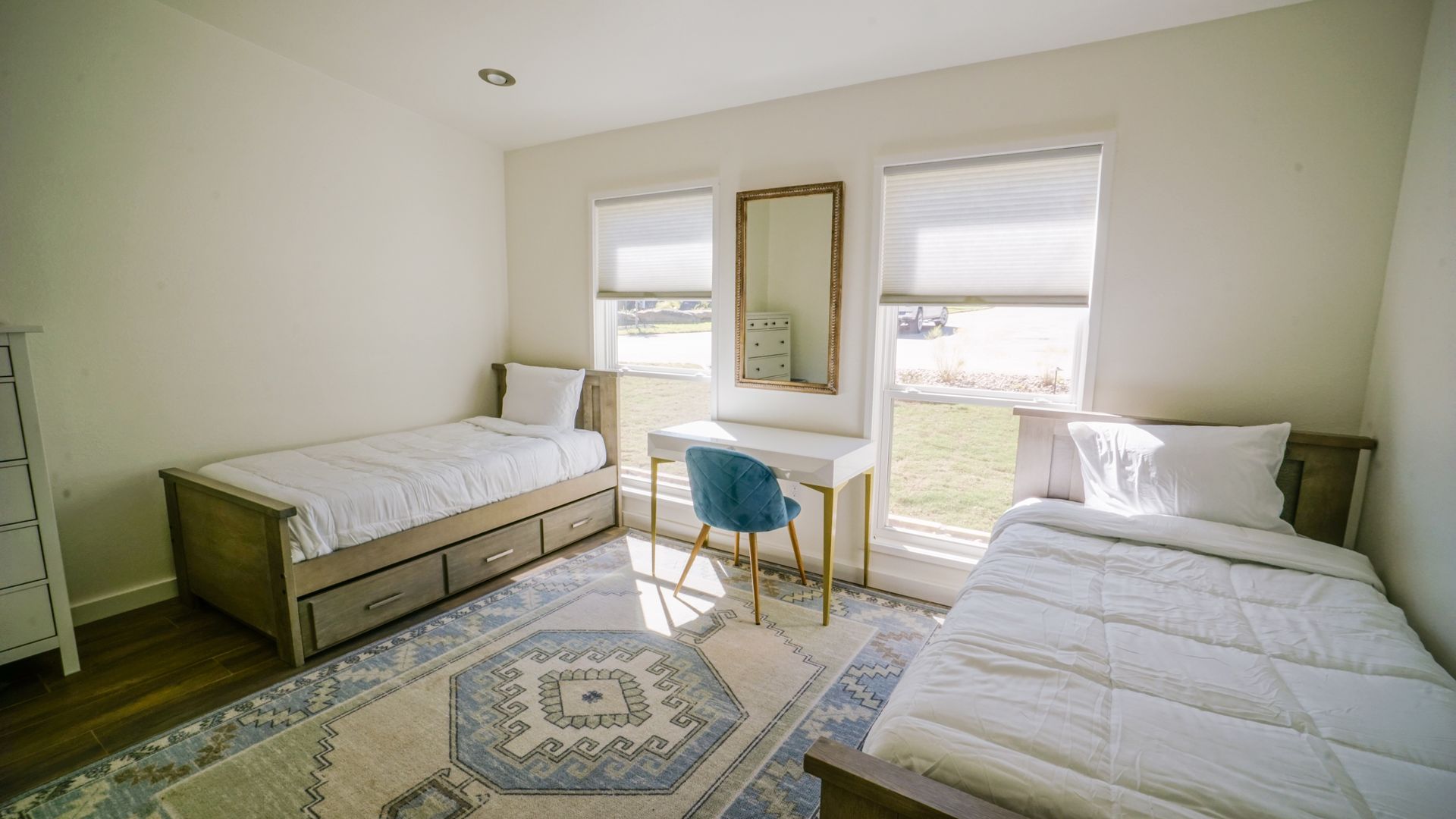 Bedroom with two beds, desk, and rug. Cream walls, blue chair, and natural light from the windows.